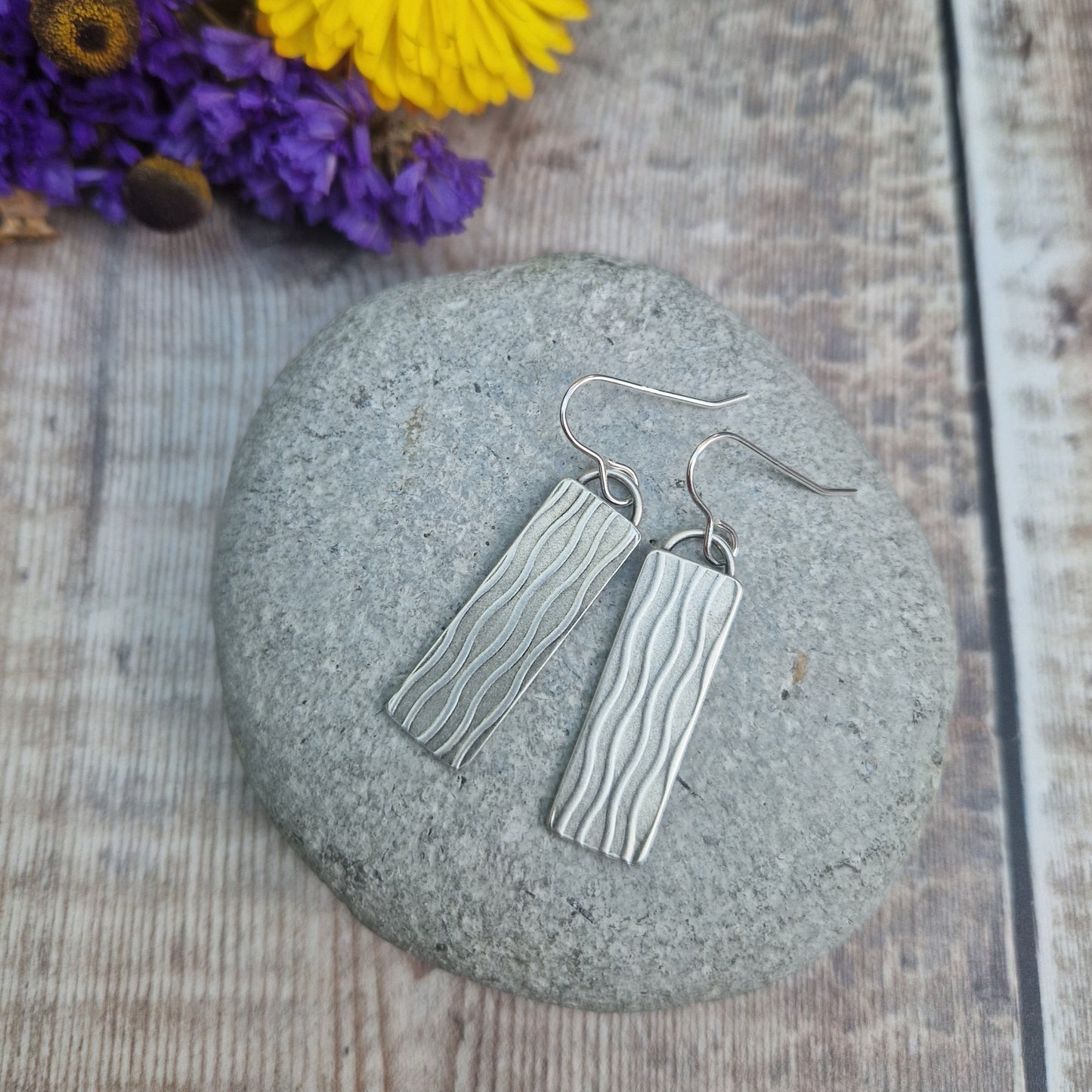 Each earring is an Oxidised Sterling Silver rectangular shaped pendant suspended from silver earring wire. Stamped on the rectangular shape are vertical wavy lines going the length of the pendant. Parts of the pattern are darker and lighter.