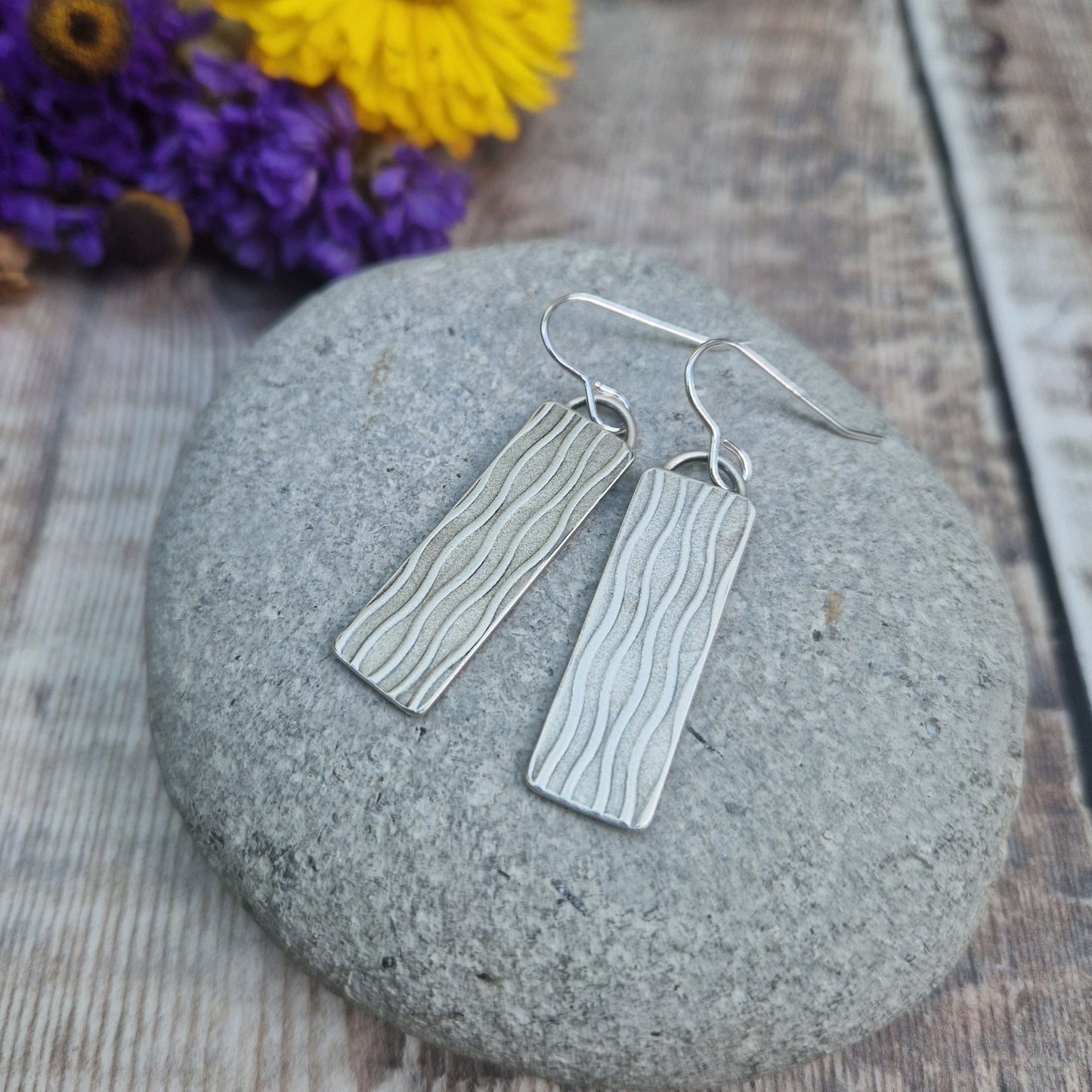 Each earring is an Oxidised Sterling Silver rectangular shaped pendant suspended from silver earring wire. Stamped on the rectangular shape are vertical wavy lines going the length of the pendant. Parts of the pattern are darker and lighter.