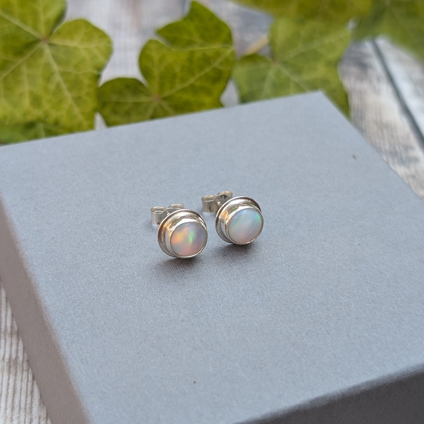 White aurora opal stud earrings handmade from recycled Sterling Silver on a grey jewellery box, showing their simple round design.