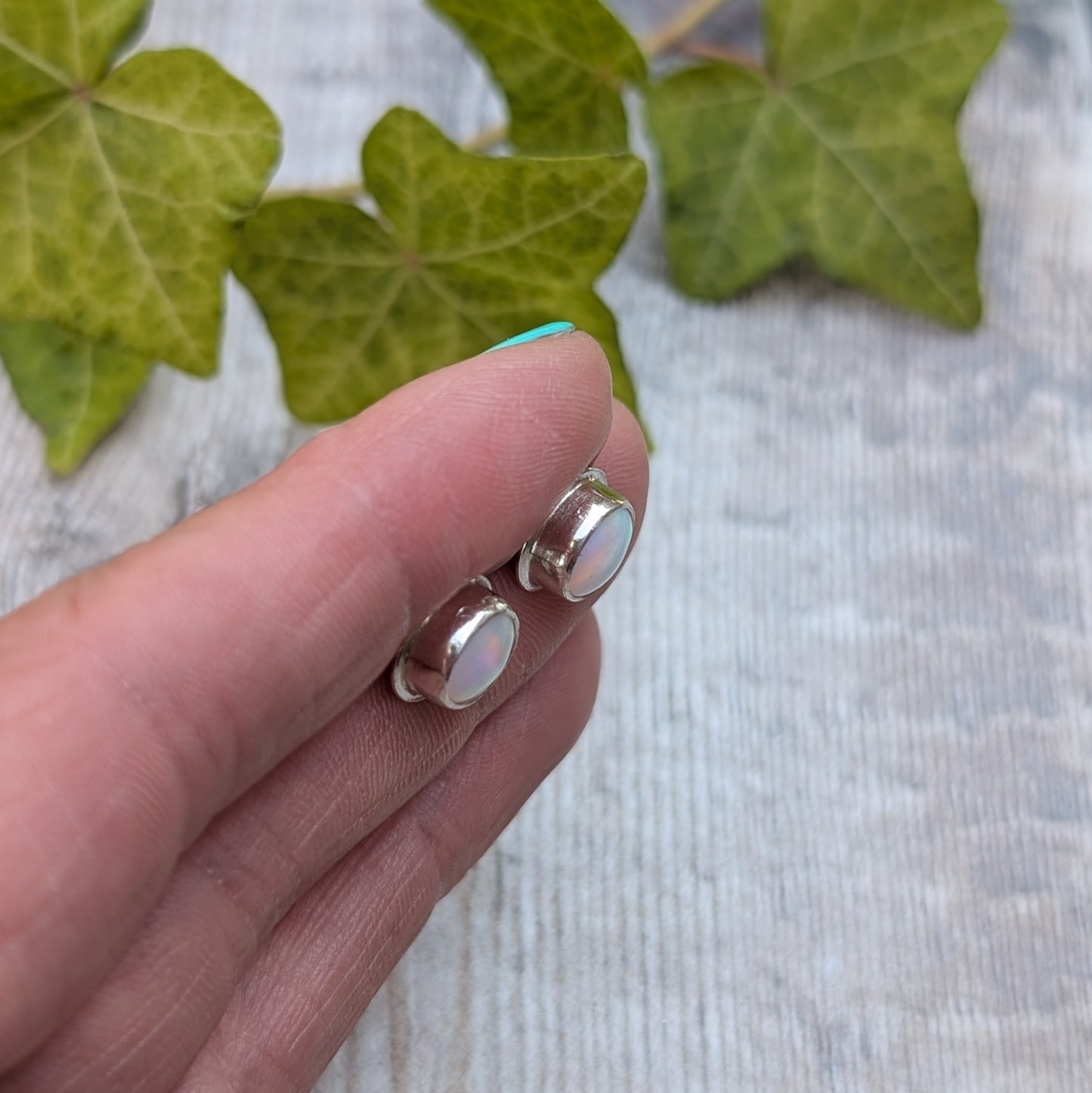 White aurora opal stud earrings handmade from recycled Sterling Silver held between fingers, showing the smooth bezel setting and colourful opals.