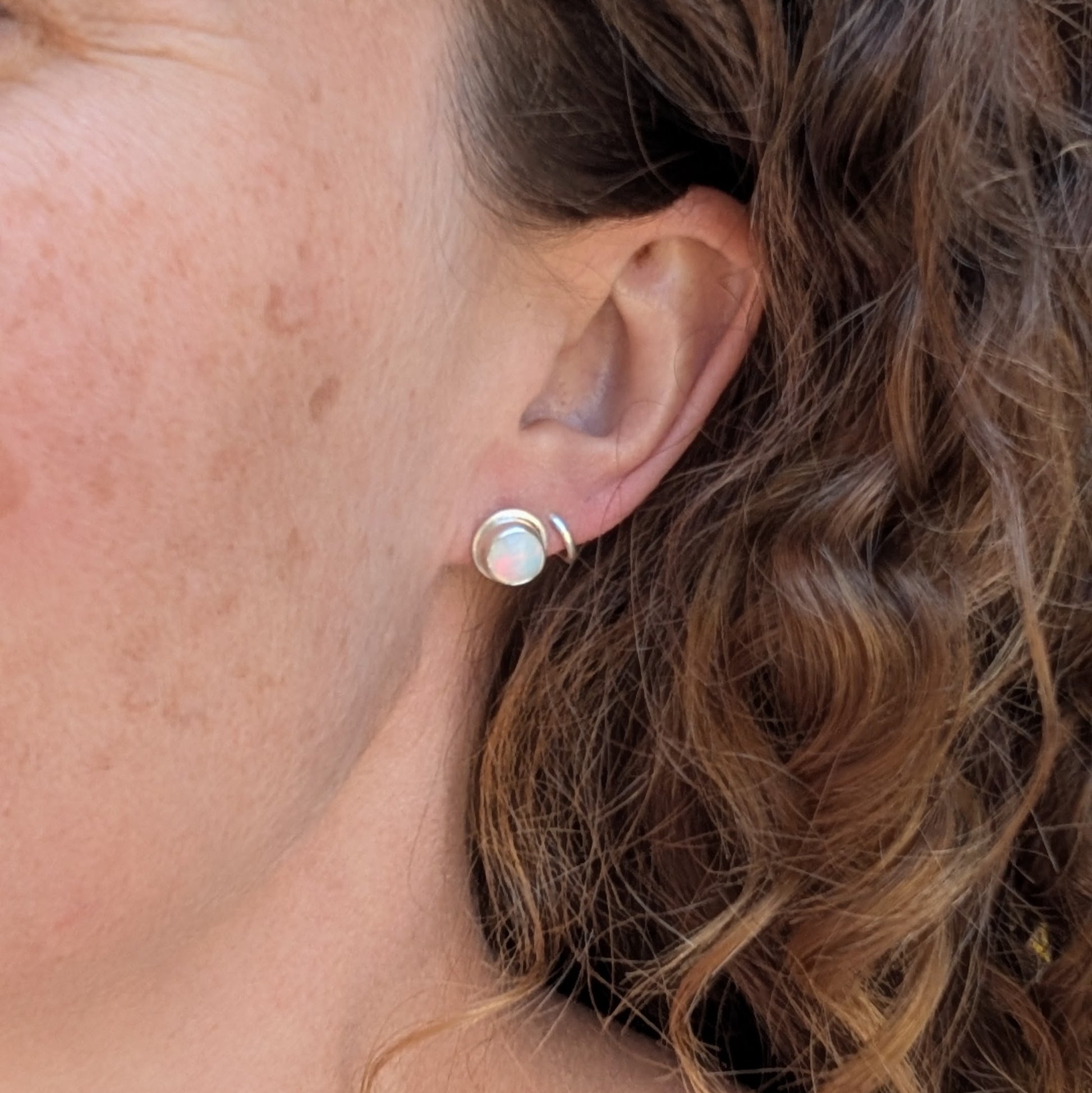 White aurora opal stud earrings handmade from recycled Sterling Silver worn in the ear, showing their small everyday size.