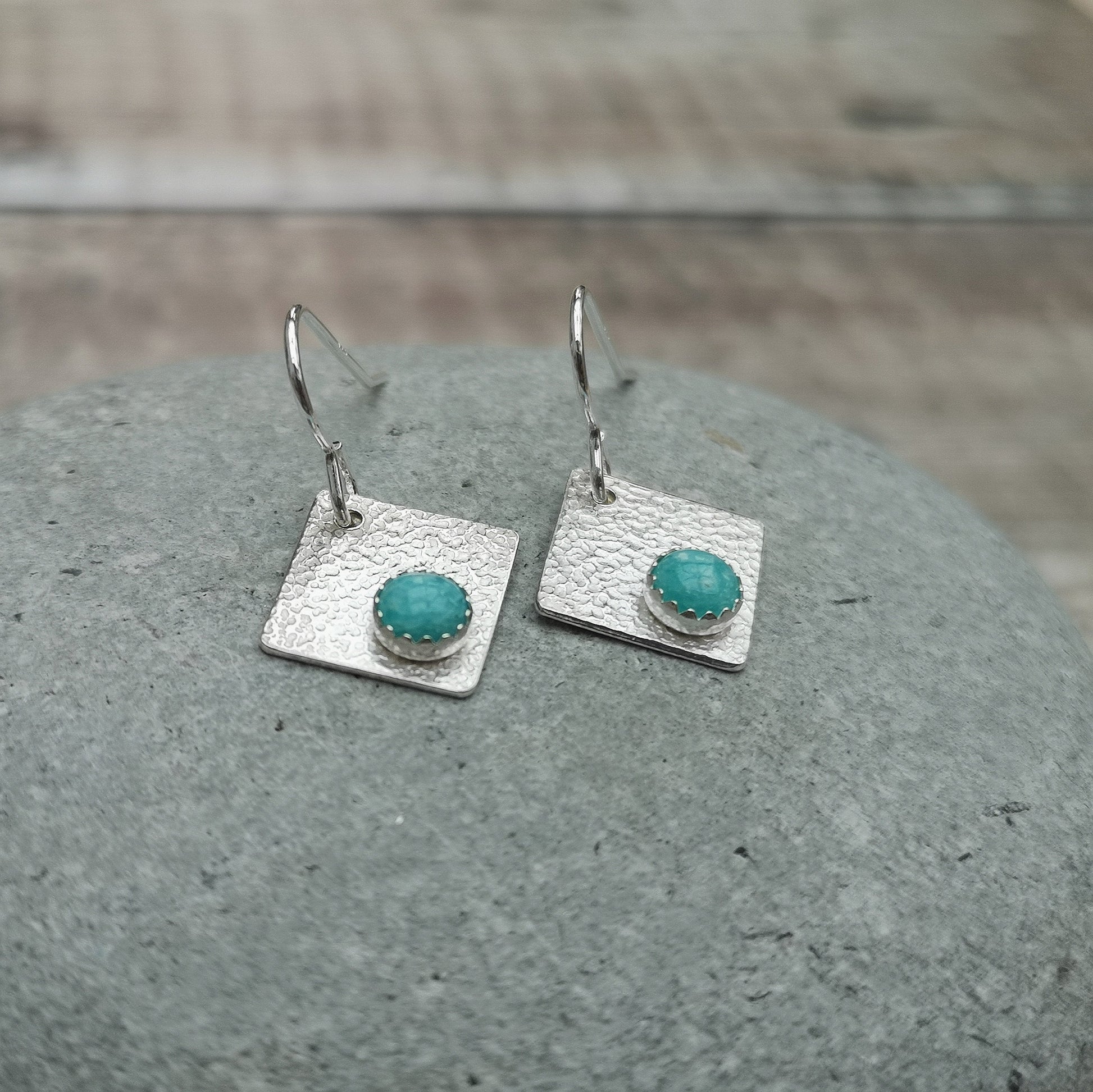 A pair of textured silver earrings featuring square panels with round Amazonite stones, displayed on a smooth stone surface.