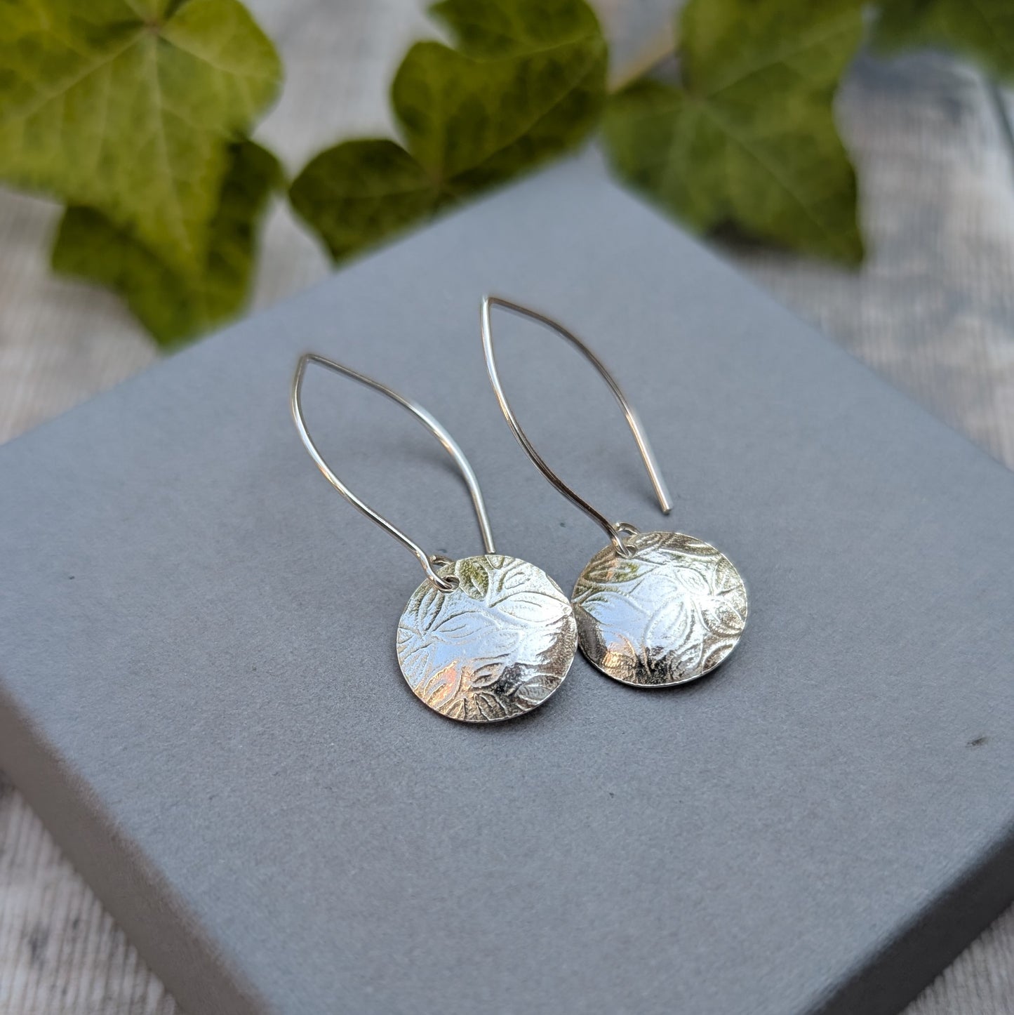 Leaf pattern disc drop earrings handmade from recycled Sterling Silver displayed on a grey jewellery box with a soft natural background.