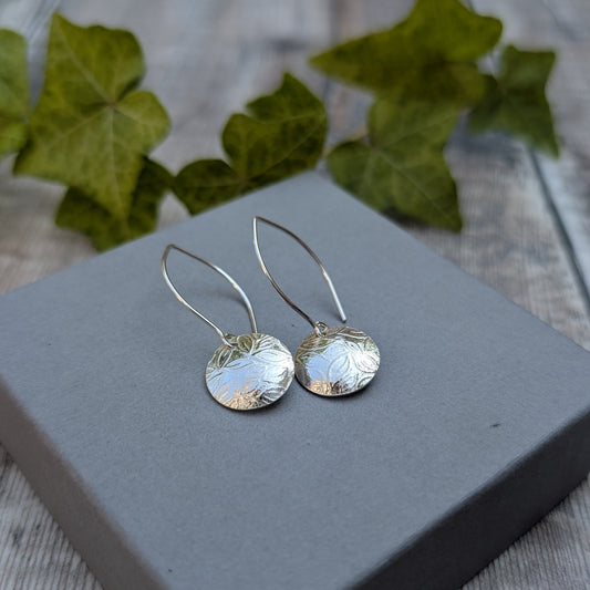 Leaf pattern disc drop earrings handmade from recycled Sterling Silver displayed on a grey jewellery box with green ivy in the background.
