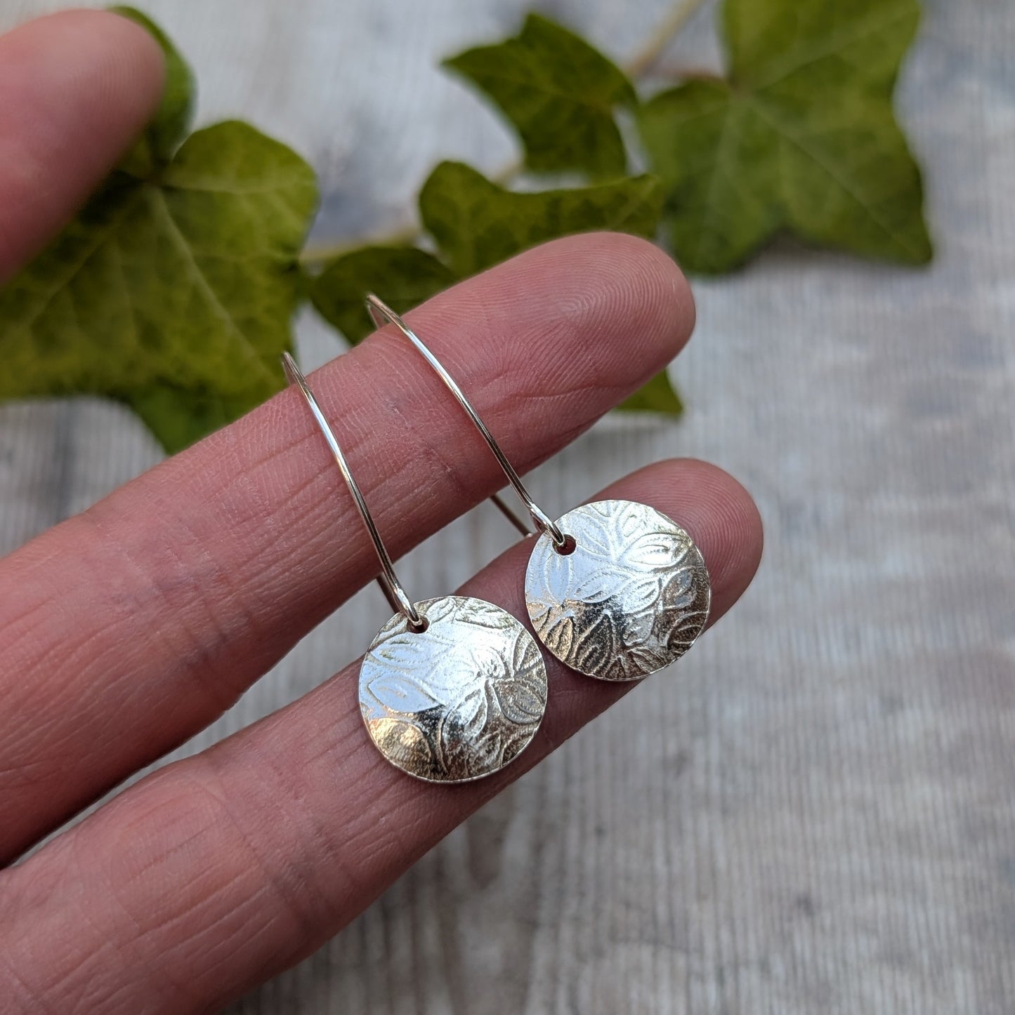Leaf pattern disc drop earrings handmade from recycled Sterling Silver held on fingertips to show their size and textured finish.