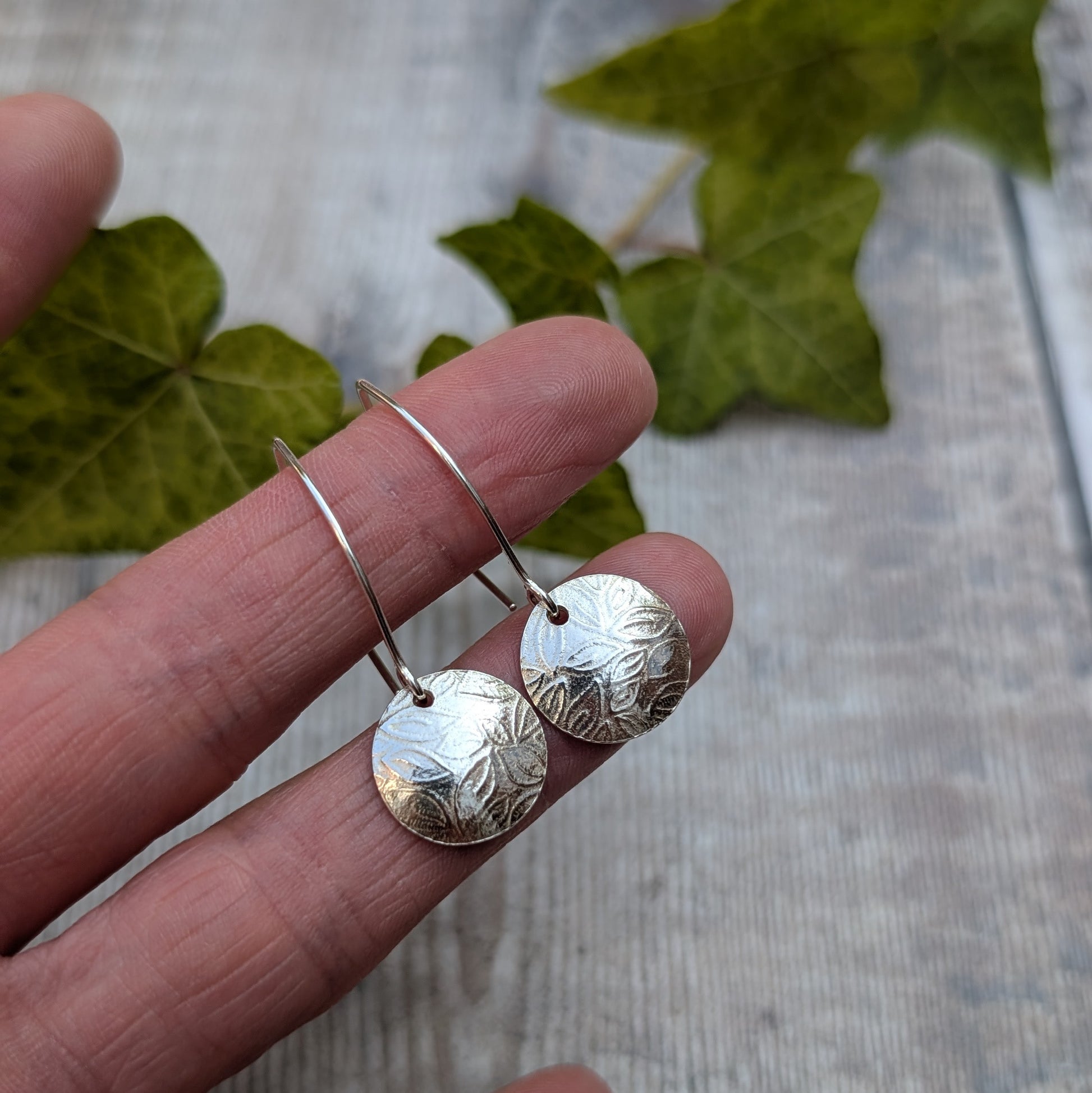 Leaf pattern disc drop earrings handmade from recycled Sterling Silver displayed on fingertips with a leafy background.
