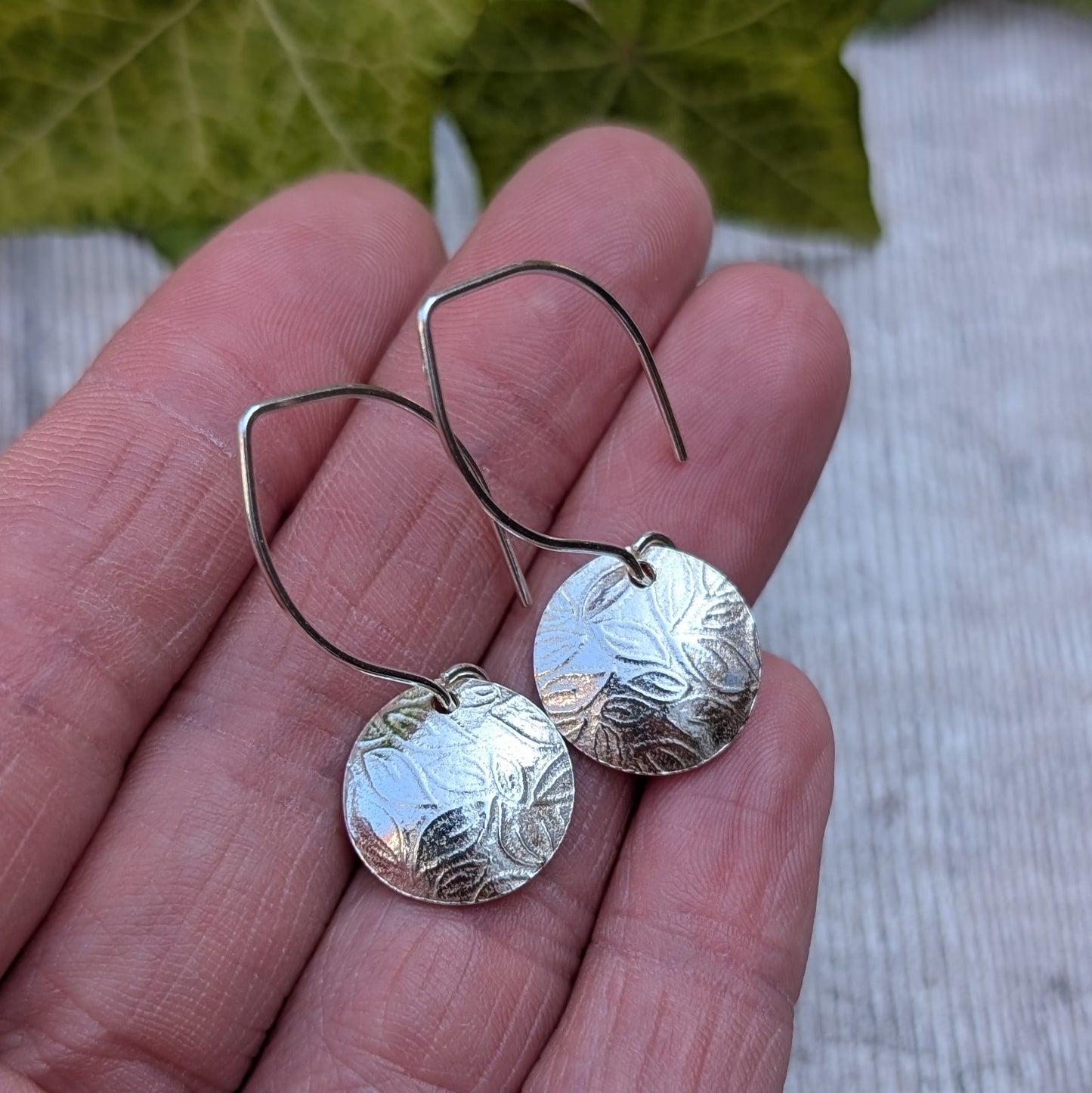 Back view of leaf pattern disc drop earrings handmade from recycled Sterling Silver resting on a hand, showing the ear wires and reverse of the discs.
