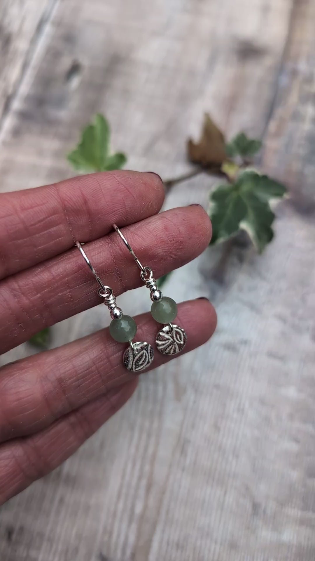Video of Close up of green aventurine and silver leaf earrings held in a hand, showing the detailed leaf pattern on the recycled sterling silver discs and soft green aventurine beads.