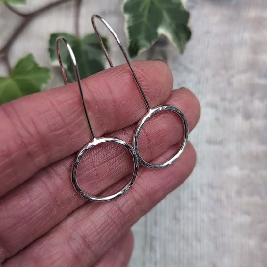 Video of handmade silver hammered circle long earrings held against a hand, showing the shiny hammered texture, open circle design and long silver ear wires, with ivy leaves in the background.