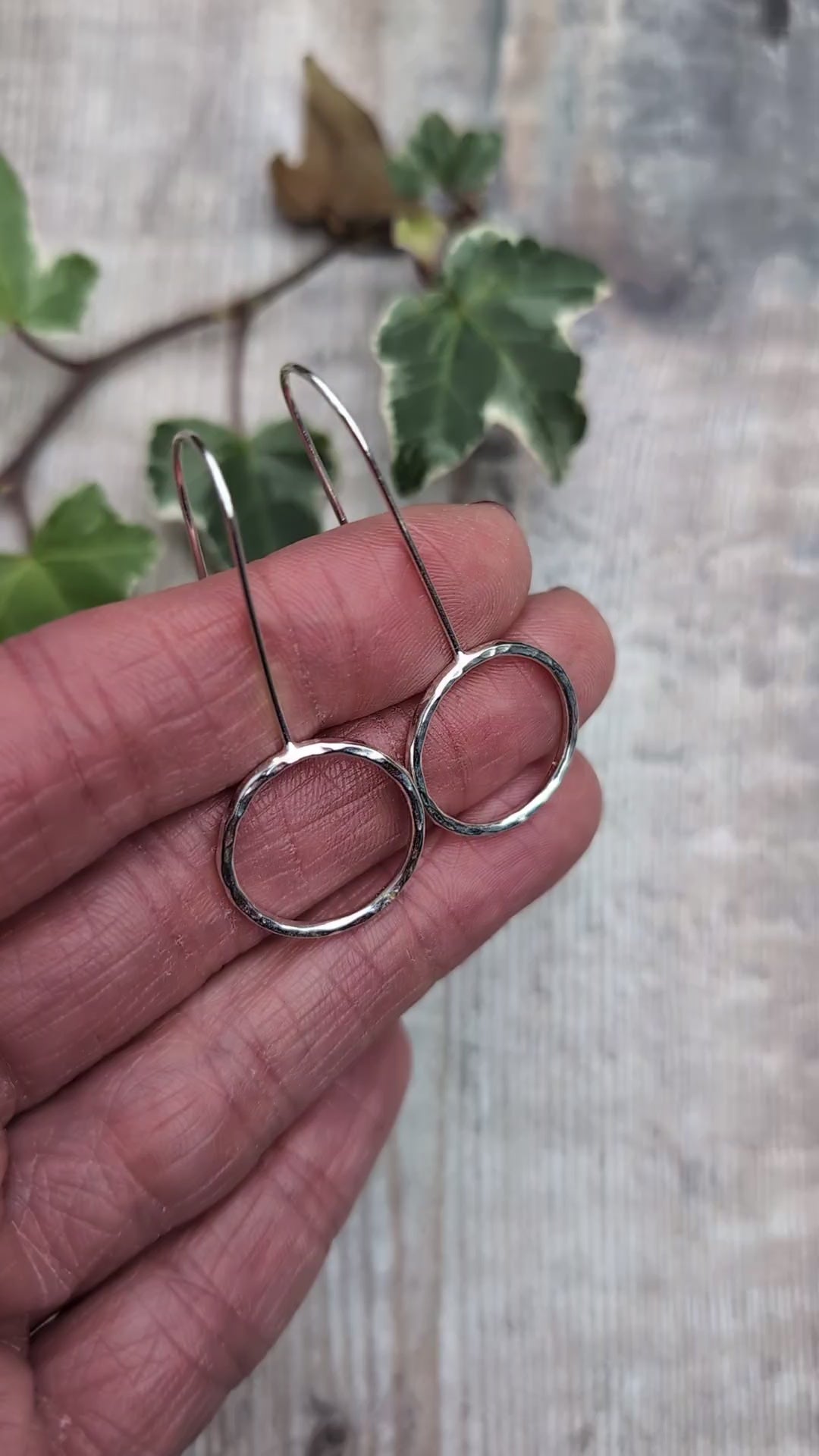 Video of handmade silver hammered circle long earrings held against a hand, showing the shiny hammered texture, open circle design and long silver ear wires, with ivy leaves in the background.