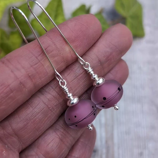Video of pink scribbles lampwork glass long silver earrings held in a hand, showing the frosted pink beads with black scribble and dot patterns.