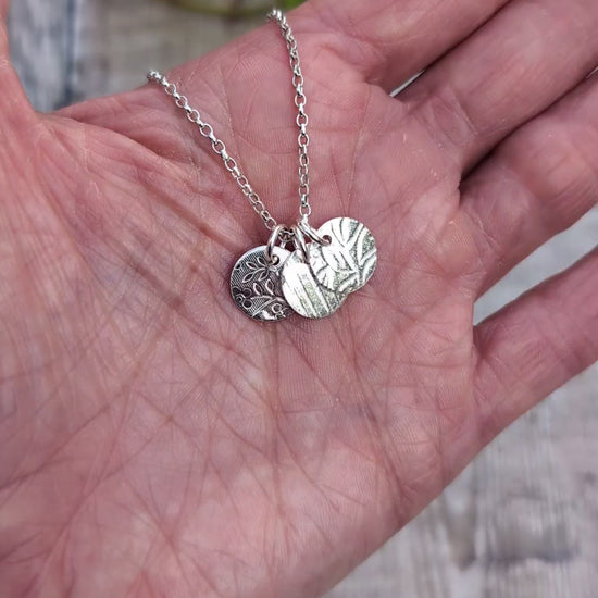 Video of Silver wildflower trio of discs necklace handmade from recycled Sterling Silver, shown resting in a hand with three different patterned silver discs.