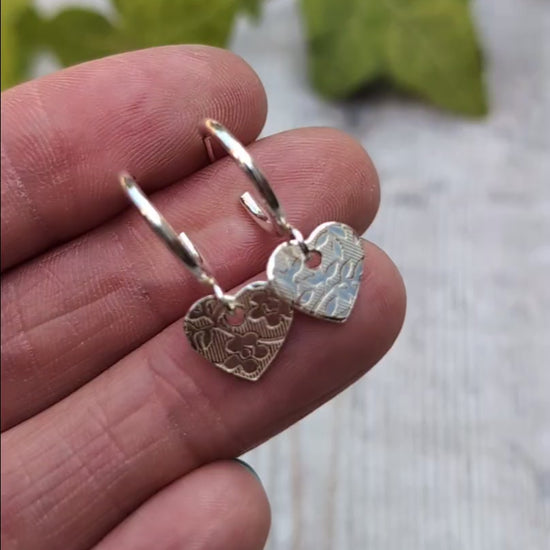 Close up of silver heart drop hoop earrings handmade from recycled Sterling Silver held in the hand showing the floral textured heart charms.