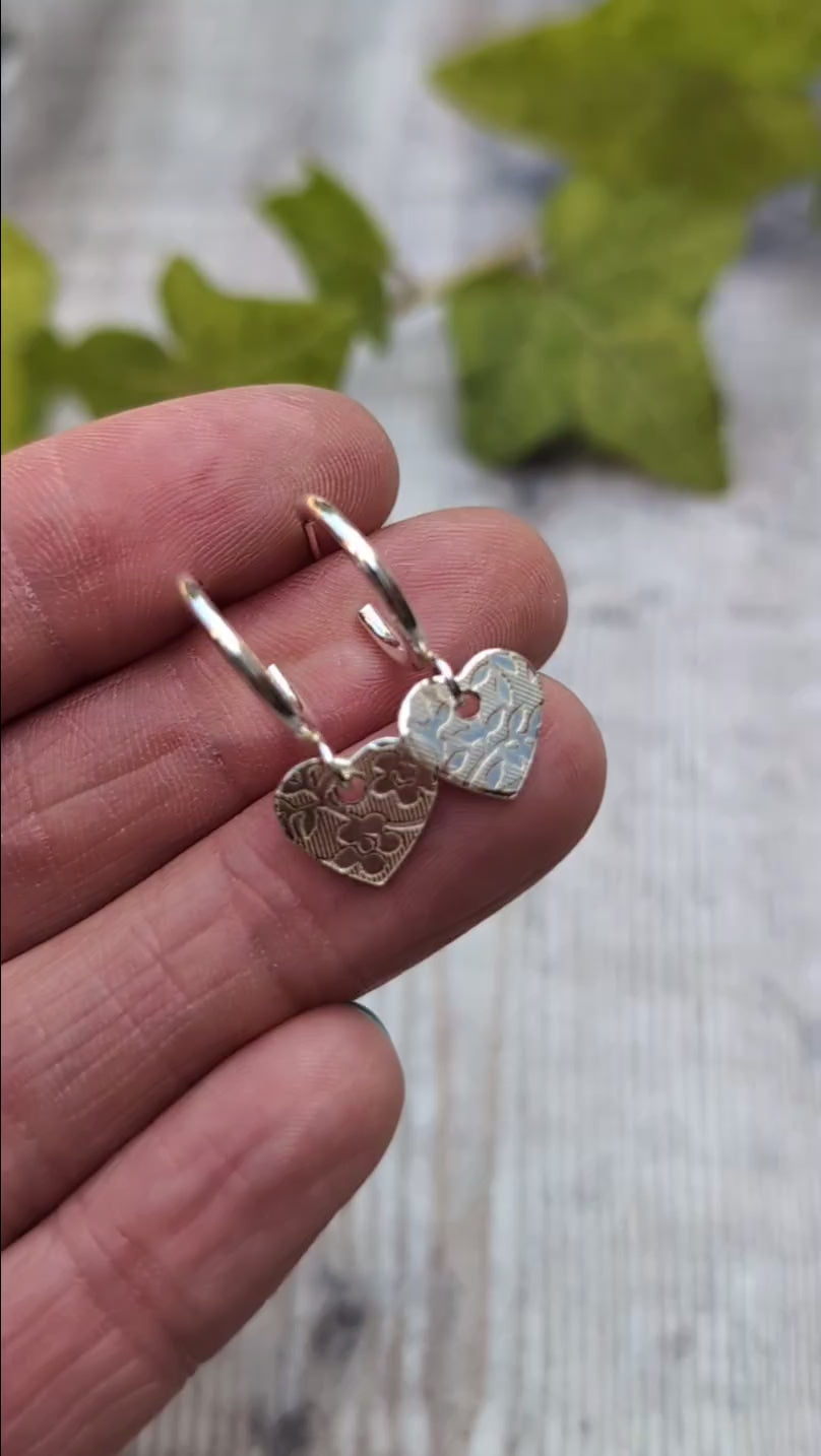 Close up of silver heart drop hoop earrings handmade from recycled Sterling Silver held in the hand showing the floral textured heart charms.