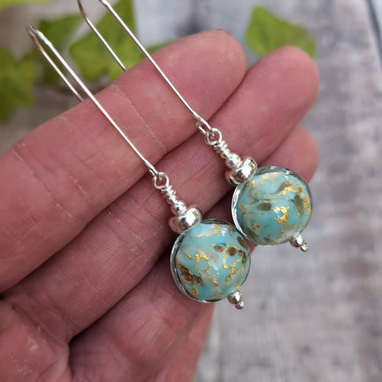 Video of Close up of turquoise marbled Venetian glass long silver earrings held in a hand, with turquoise blue and gold handmade lampwork glass beads.
