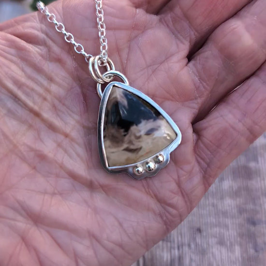 Video of Petrified Palm Wood Statement Gemstone Necklace resting in the palm of a hand, showing the natural patterns of the triangular gemstone, polished silver setting and three decorative silver balls.