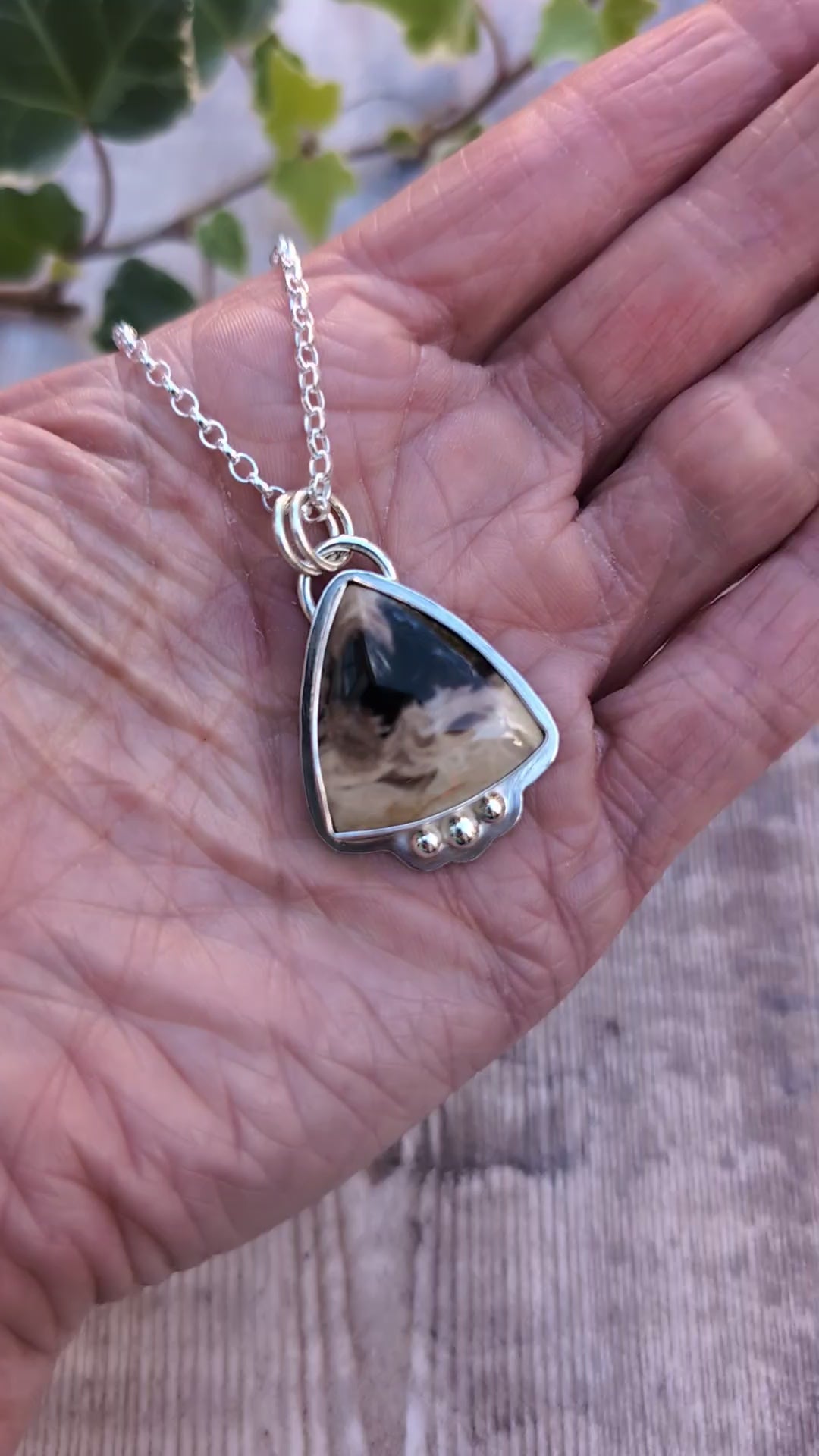 Video of Petrified Palm Wood Statement Gemstone Necklace resting in the palm of a hand, showing the natural patterns of the triangular gemstone, polished silver setting and three decorative silver balls.