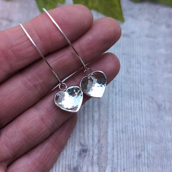 Hammered silver heart long earrings handmade from recycled Sterling Silver, close up on a hand showing the hammered heart detail and polished finish.