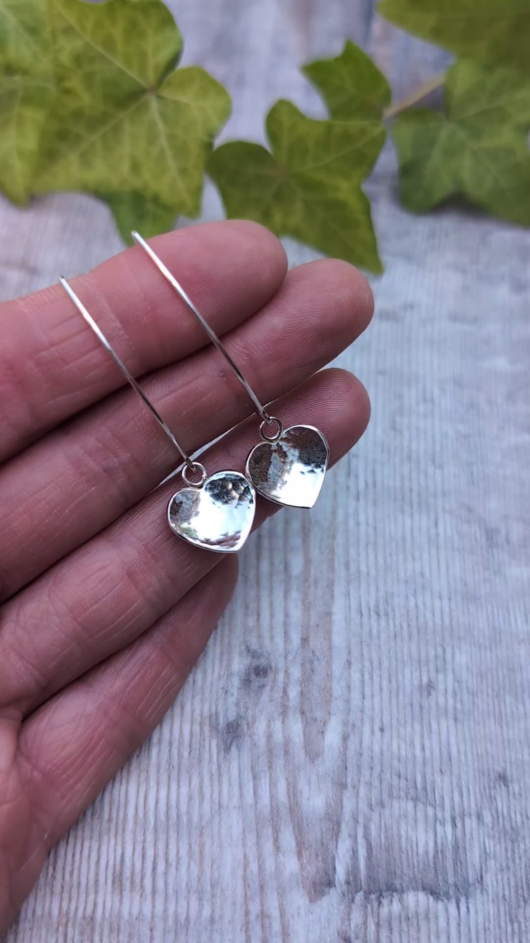 Hammered silver heart long earrings handmade from recycled Sterling Silver, close up on a hand showing the hammered heart detail and polished finish.