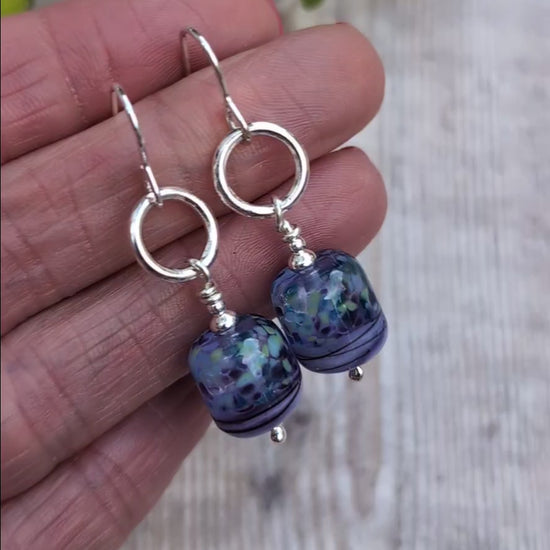 Video of Lilac Watercolour Lampwork Glass and Silver Circle Earrings held in a hand, showing the lilac, blue and green pattern of the handmade Lampwork glass beads.