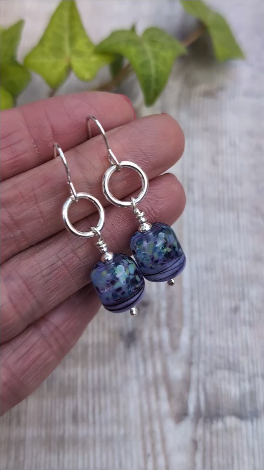 Video of Lilac Watercolour Lampwork Glass and Silver Circle Earrings held in a hand, showing the lilac, blue and green pattern of the handmade Lampwork glass beads.