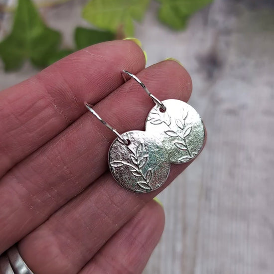 Video of Silver leafy vine oval disc earrings held in a hand, showing the pressed leafy vine pattern and handmade silver ear wires.