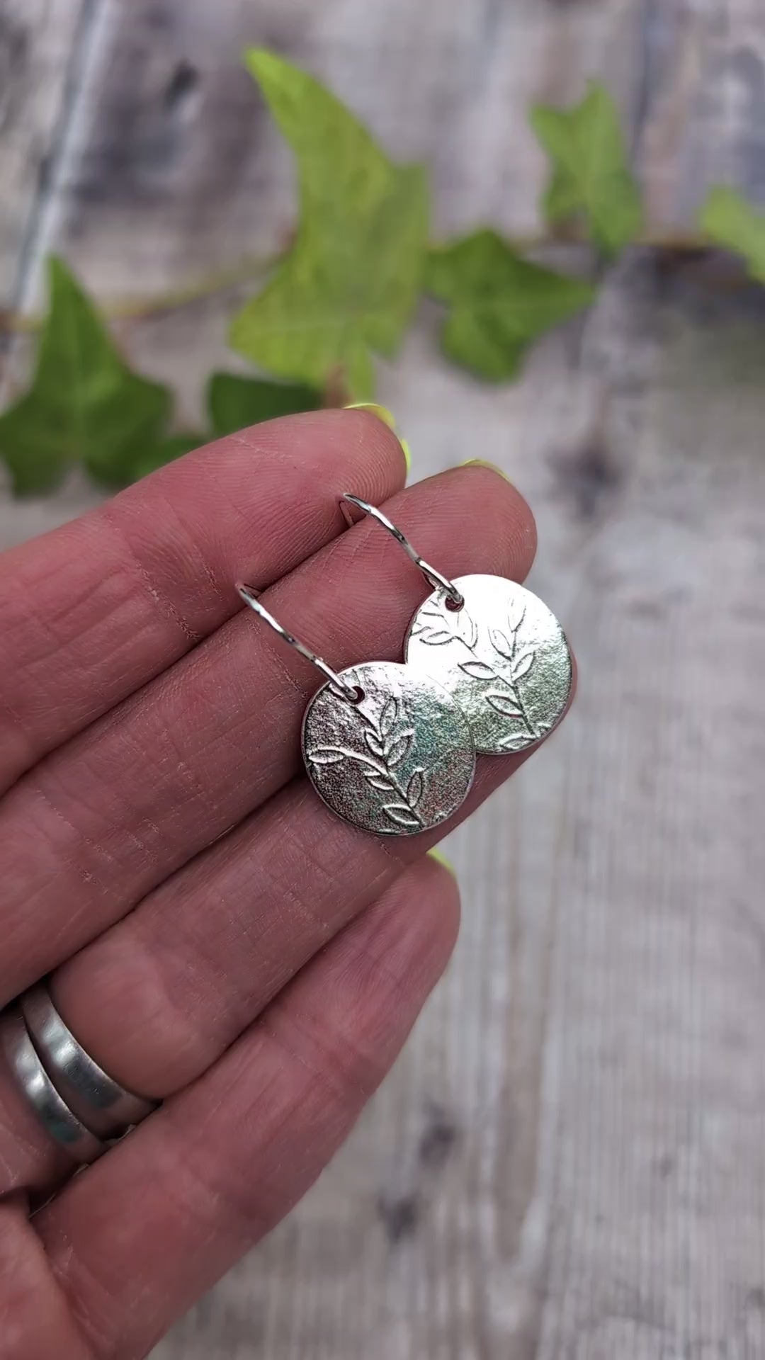 Video of Silver leafy vine oval disc earrings held in a hand, showing the pressed leafy vine pattern and handmade silver ear wires.