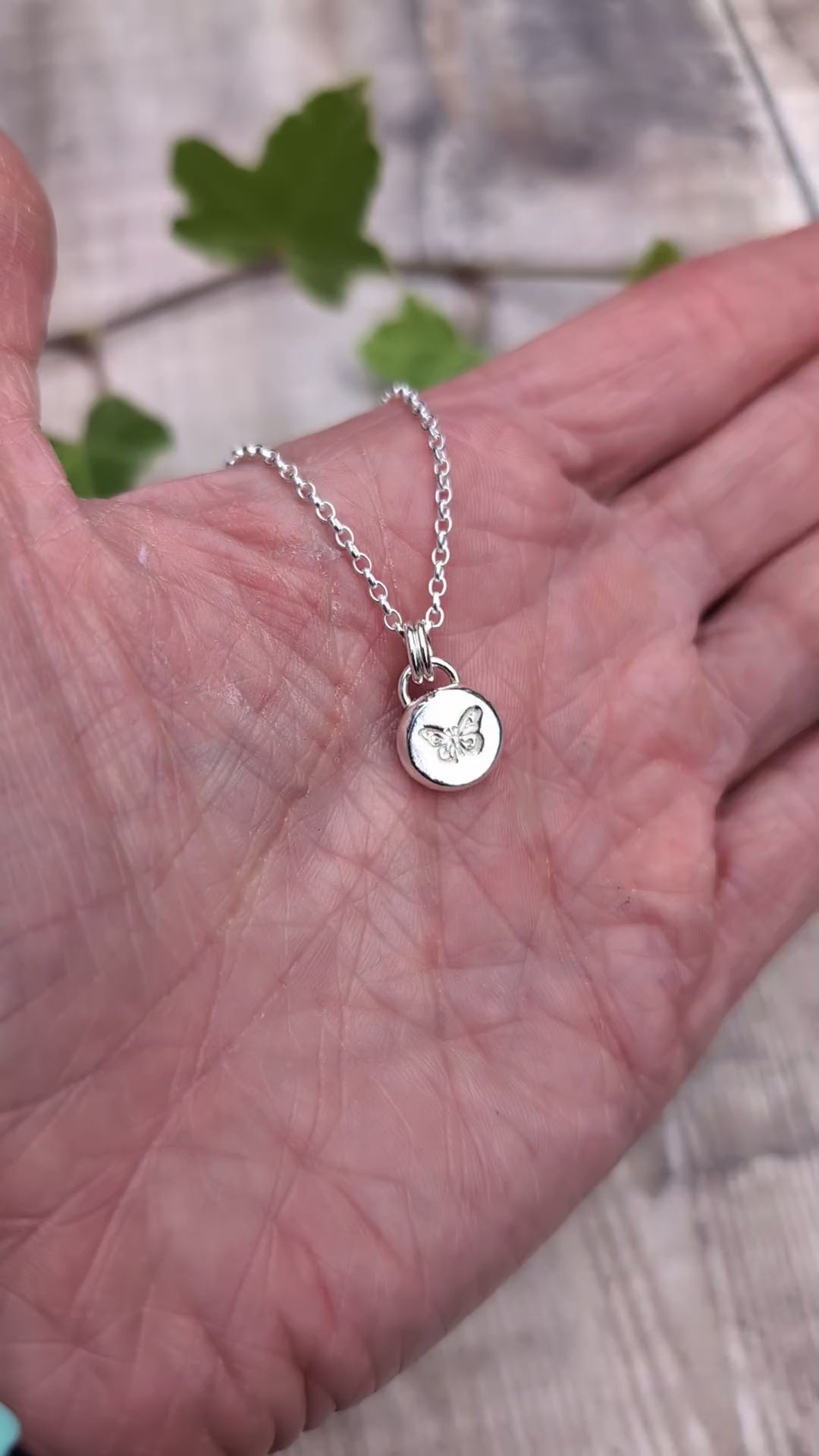 Video of Silver butterfly pebble necklace held in a hand, showing the size and hand stamped butterfly design on the small silver pendant.