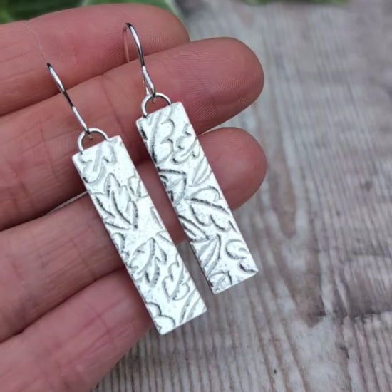 Video of silver autumn leaves long rectangle earrings held in a hand, showing the detailed autumn leaf pattern created using a rolling mill.