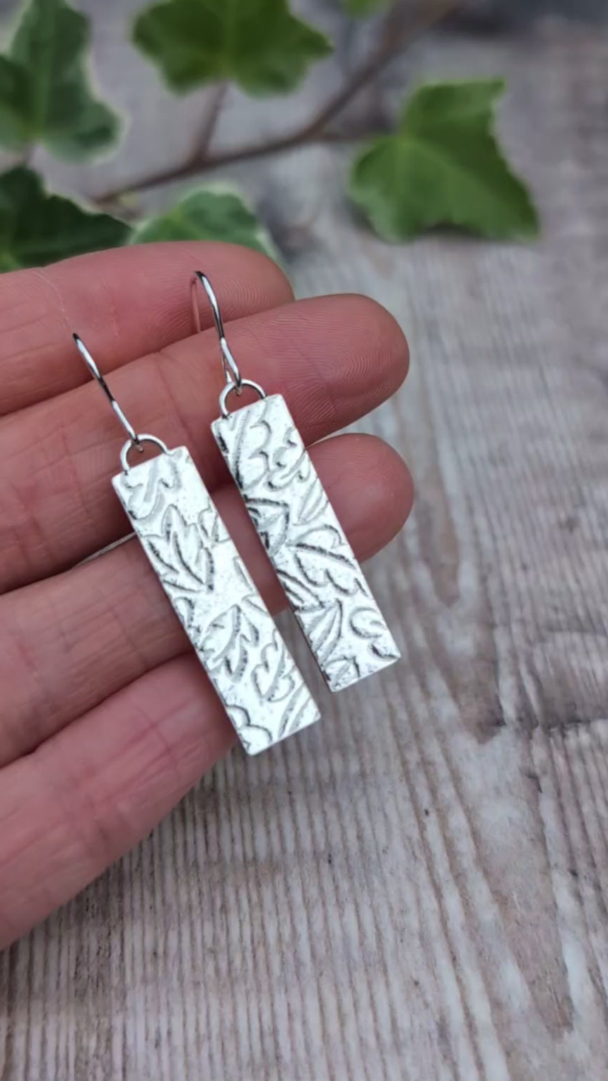 Video of silver autumn leaves long rectangle earrings held in a hand, showing the detailed autumn leaf pattern created using a rolling mill.