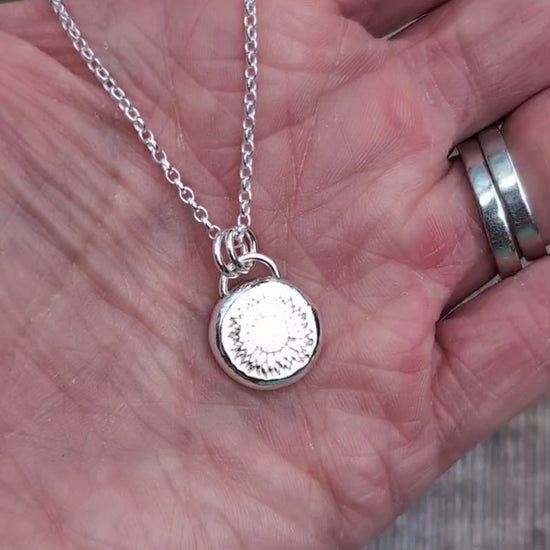 Video of Close up of the Silver Sunflower Pebble Necklace held in a hand, showing the detailed sunflower stamped into the round silver pebble pendant.
