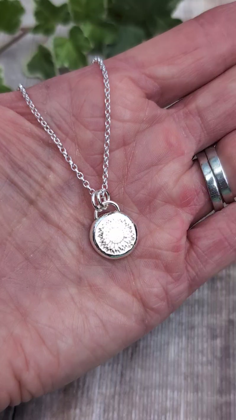Video of Close up of the Silver Sunflower Pebble Necklace held in a hand, showing the detailed sunflower stamped into the round silver pebble pendant.