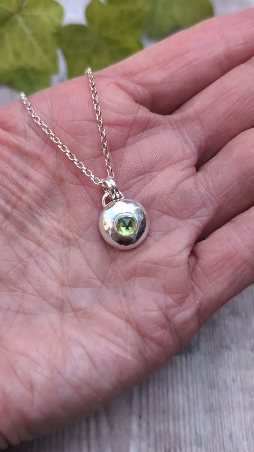 Peridot pebble necklace resting in the palm of a hand, showing the size of the handmade recycled Sterling Silver pebble pendant with a faceted Peridot gemstone.