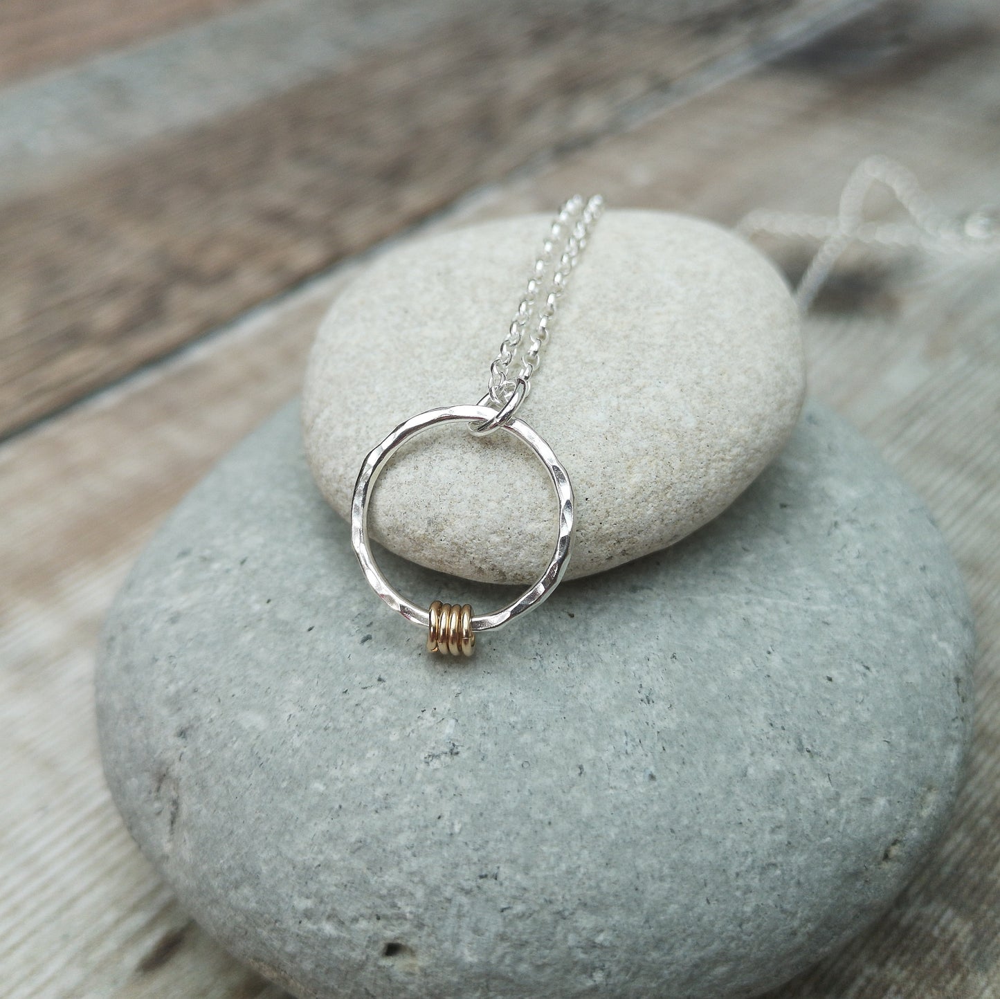 A silver hammered circle pendant necklace with four small gold rings, displayed on a smooth stone surface, showcasing the elegant combination of mixed metals.