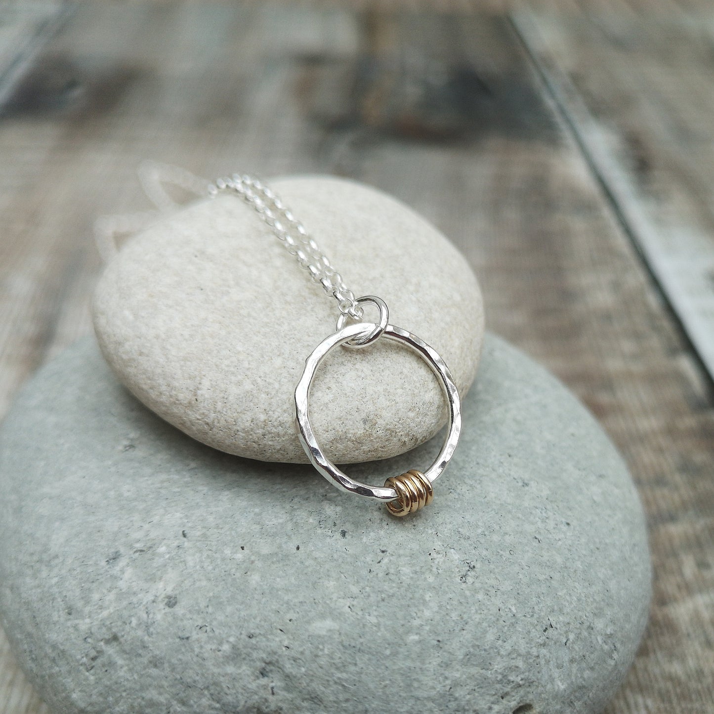 A silver hammered circle pendant necklace with four small gold rings, displayed on a smooth stone surface, showcasing the elegant combination of mixed metals.