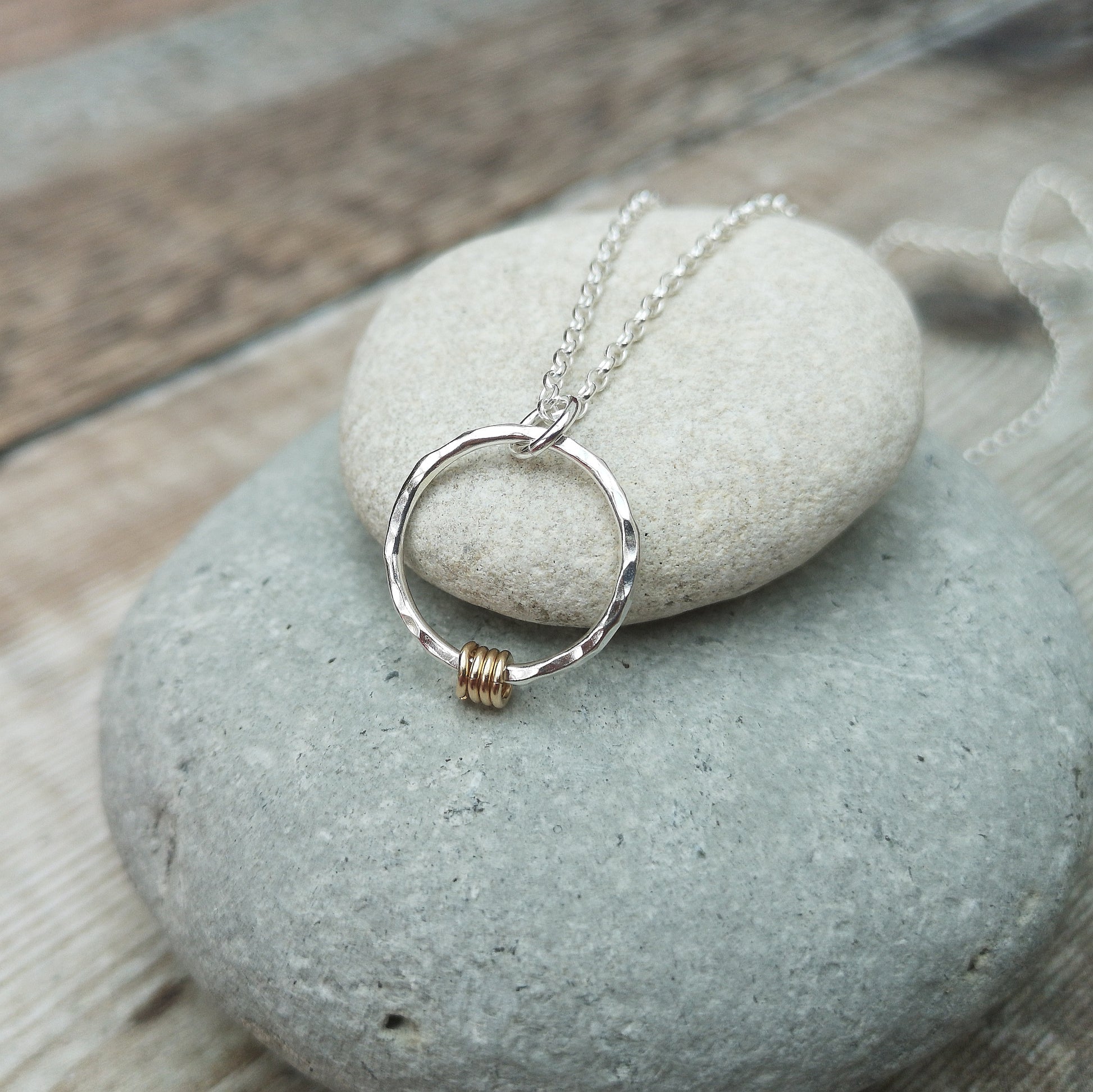 A silver hammered circle pendant necklace with four small gold rings, displayed on a smooth stone surface, showcasing the elegant combination of mixed metals.