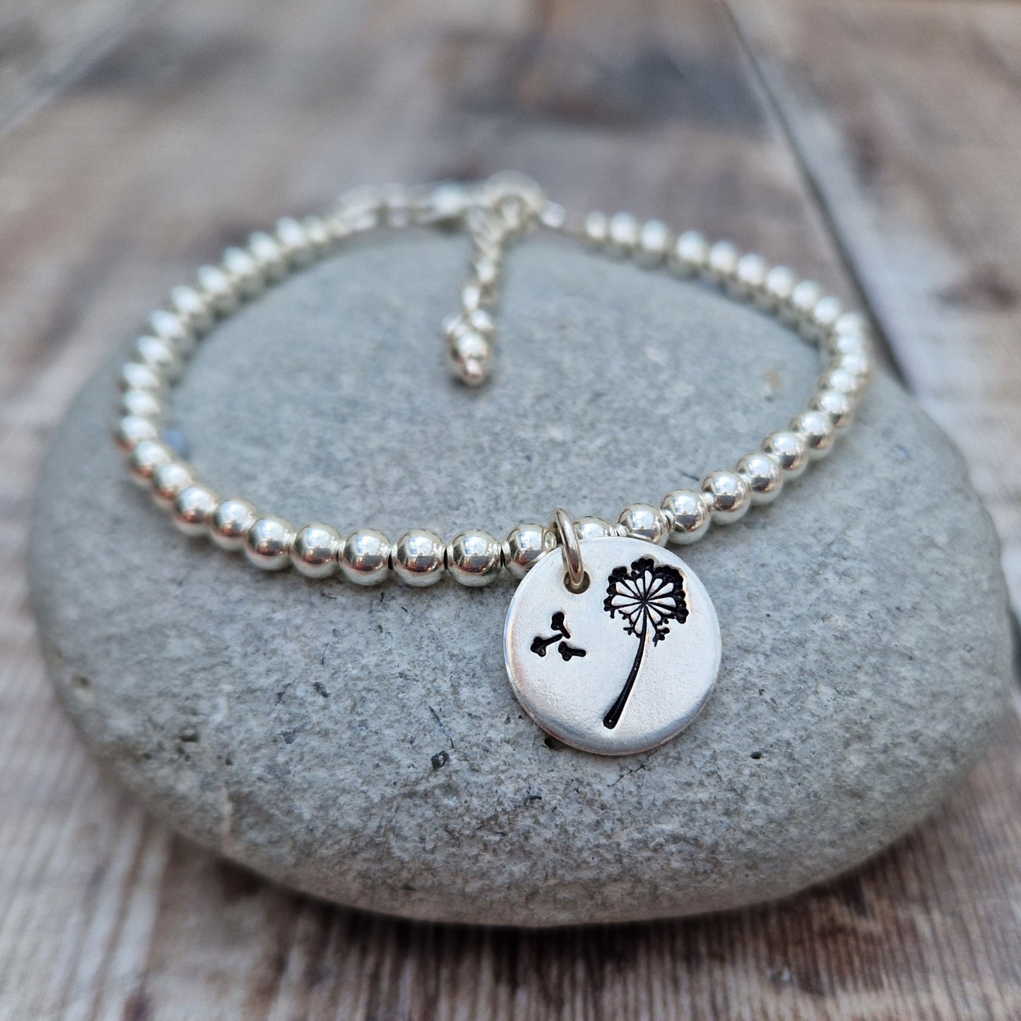 Silver beaded bracelet with a dandelion charm, displayed on a smooth stone. The charm features a dandelion with seeds blowing in the wind. Background shows a rustic wooden surface.