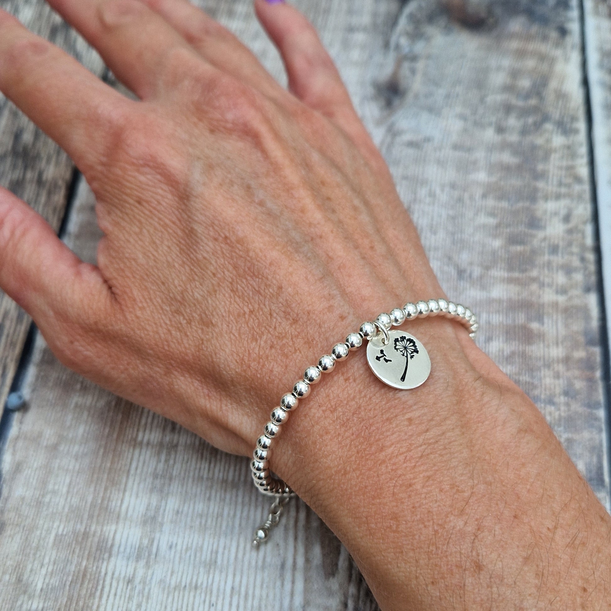 Silver beaded bracelet with a dandelion charm, worn on a wrist. The charm showcases a dandelion with seeds blowing away, symbolizing wishes and dreams. The bracelet sits comfortably against the skin.