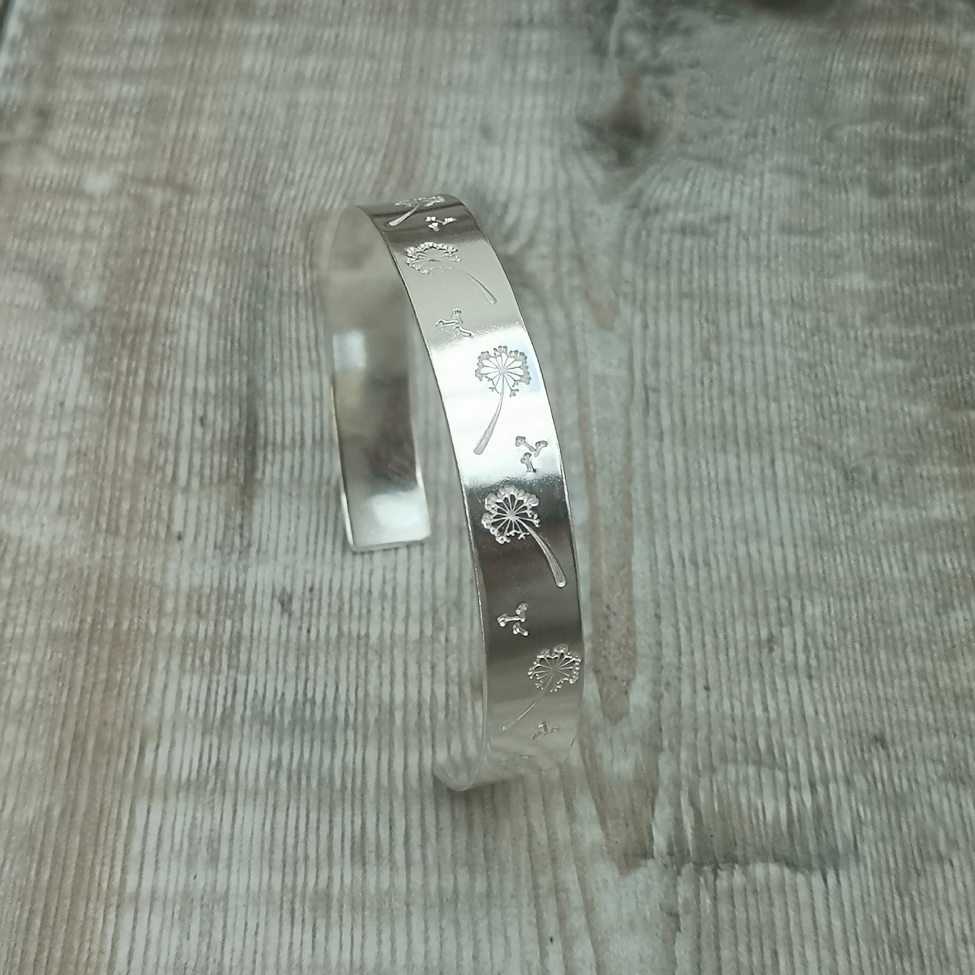 Silver cuff bangle with dandelion engravings, displayed upright on a wooden surface. The detailed engravings show dandelions and seeds blowing in the wind, creating a delicate and whimsical design