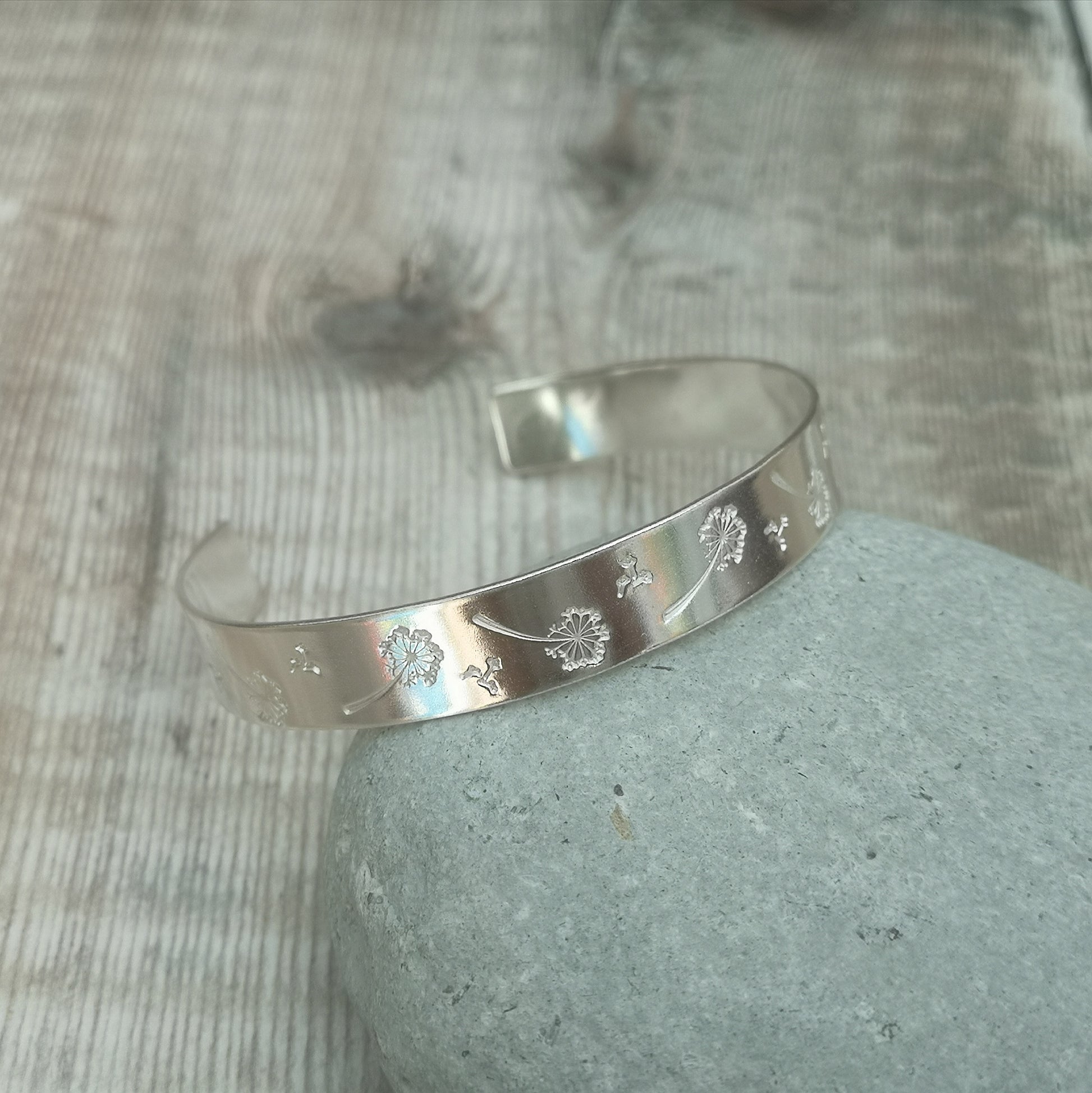 Silver cuff bangle with intricate dandelion engravings, resting on a smooth stone. The engravings depict dandelions and seeds floating away, symbolizing wishes and the fleeting nature of time.