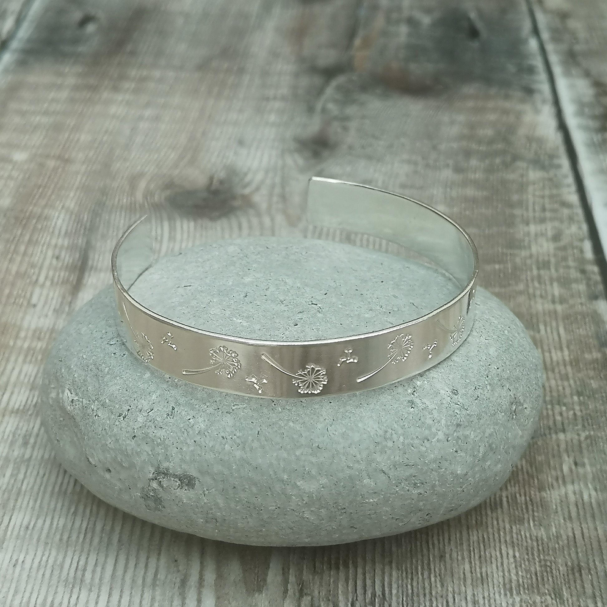 Silver cuff bangle with intricate dandelion engravings, resting on a smooth stone. The engravings depict dandelions and seeds floating away, symbolizing wishes and the fleeting nature of time.