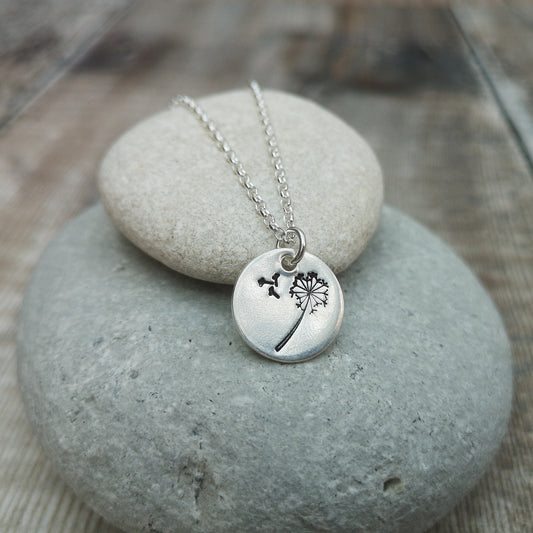 Silver necklace with a round dandelion charm, displayed on stacked smooth stones. The charm features an engraved dandelion with seeds blowing away, representing hope and wishes. The chain and charm rest gently against the stone background.