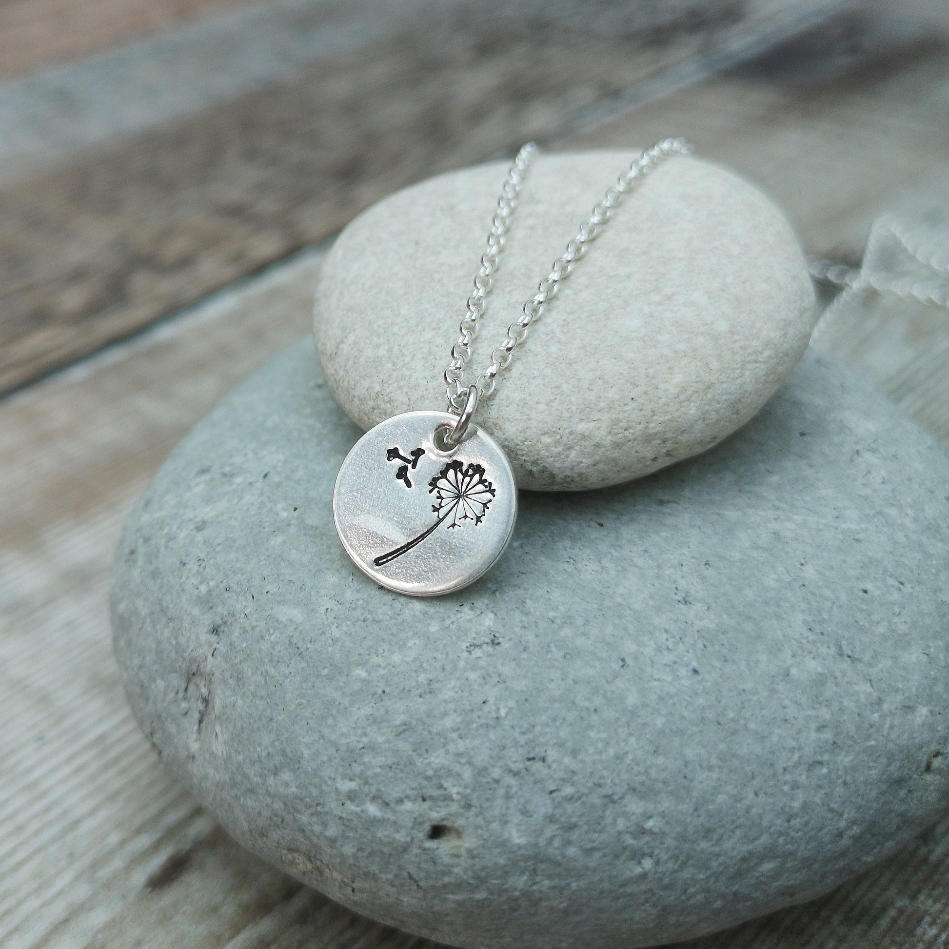 Silver necklace with a round dandelion charm, displayed on stacked smooth stones. The charm features an engraved dandelion with seeds blowing away, representing hope and wishes. The chain and charm rest gently against the stone background.