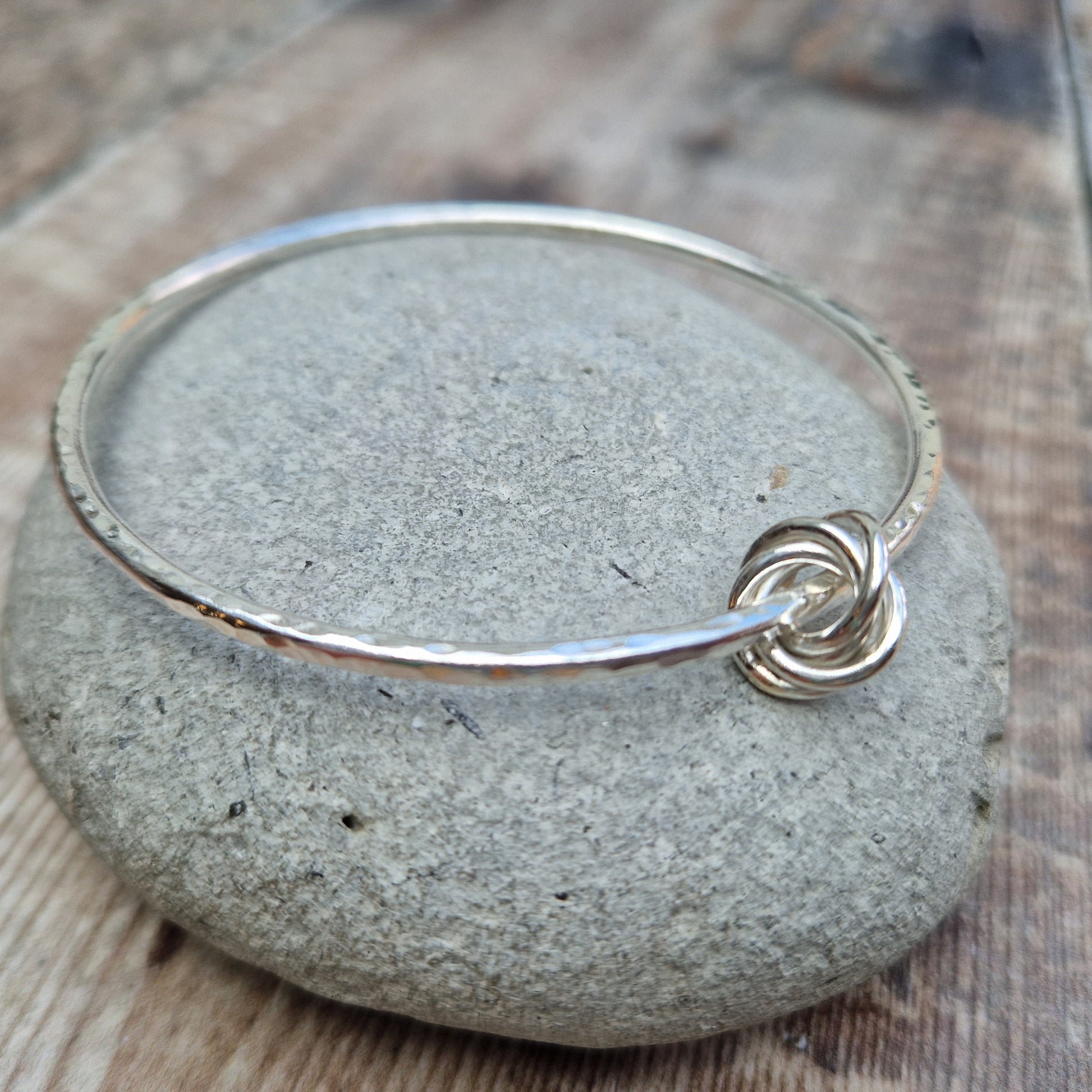 Silver hammered bangle with a knotted detail, displayed on a smooth stone. The bangle features a subtle texture, and the interlocking knot design adds a unique touch. The background includes a rustic wooden surface.