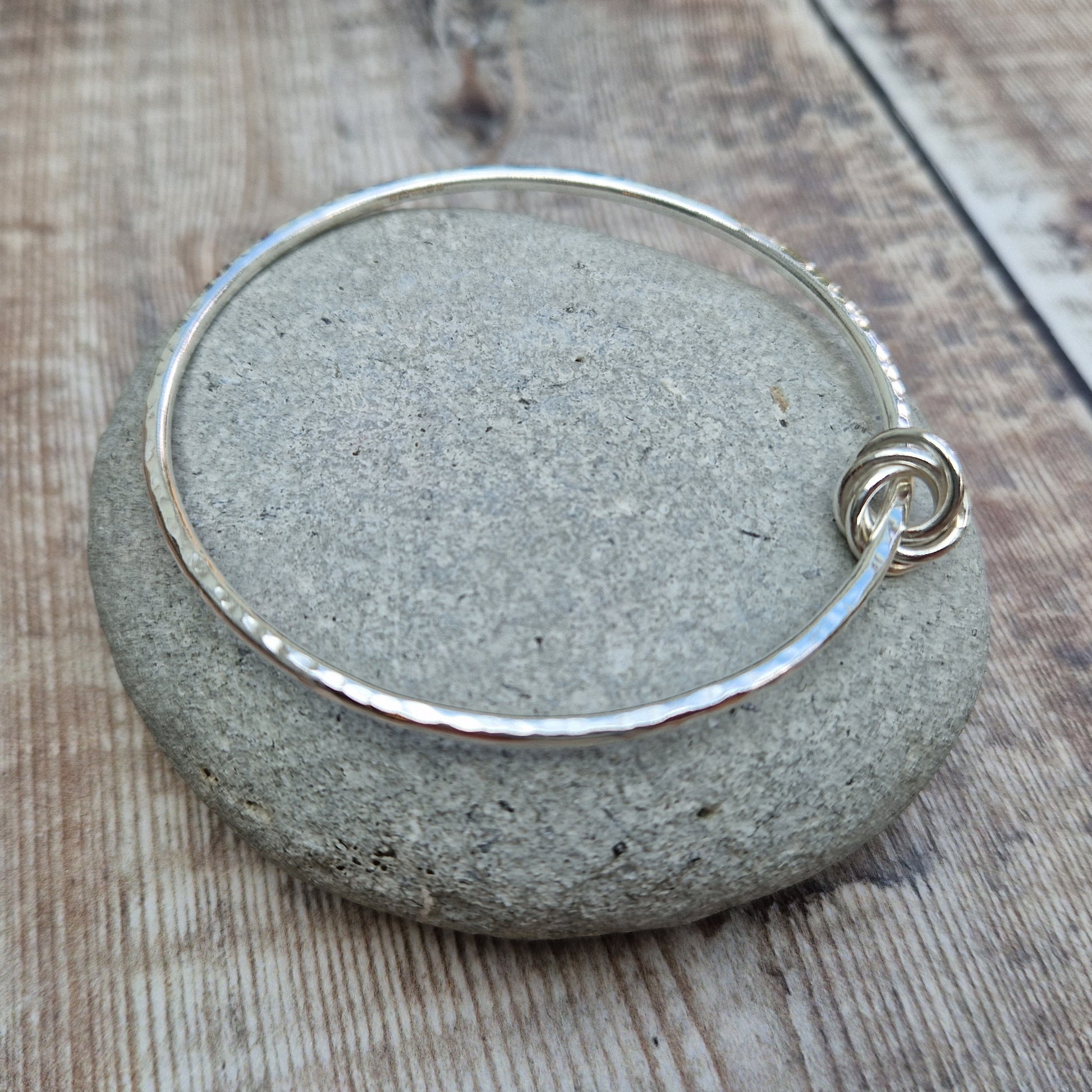 Silver hammered bangle with a knotted detail, displayed on a smooth stone. The bangle features a subtle texture, and the interlocking knot design adds a unique touch. The background includes a rustic wooden surface.