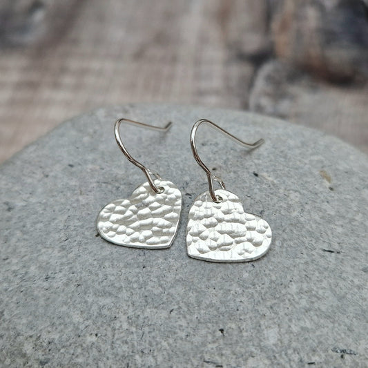 A pair of handmade sterling silver heart-shaped earrings with a hammered texture, resting on a smooth stone. The textured surface of the hearts adds depth and shimmer to the simple yet elegant design.