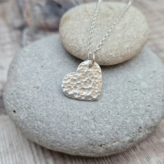 A handmade sterling silver heart pendant with a hammered texture, hanging on a delicate silver chain. The pendant is placed on a smooth stone, highlighting the simple and elegant design of the necklace.