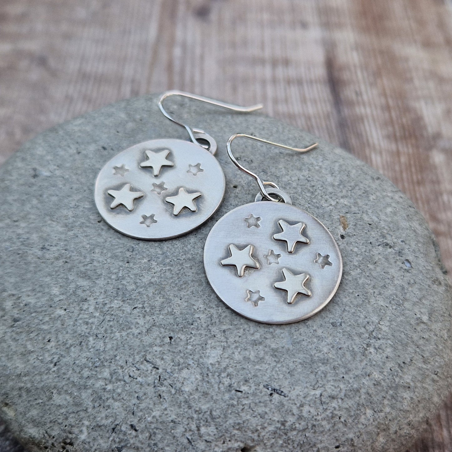 Each earring is an oxidised silver disc shape with three raised five-pointed stars and four indented smaller five-pointed stars in a random pattern on each disc. The oxidisation helps the star shapes stand out as if in shadow. Suspended from silver earring wire.