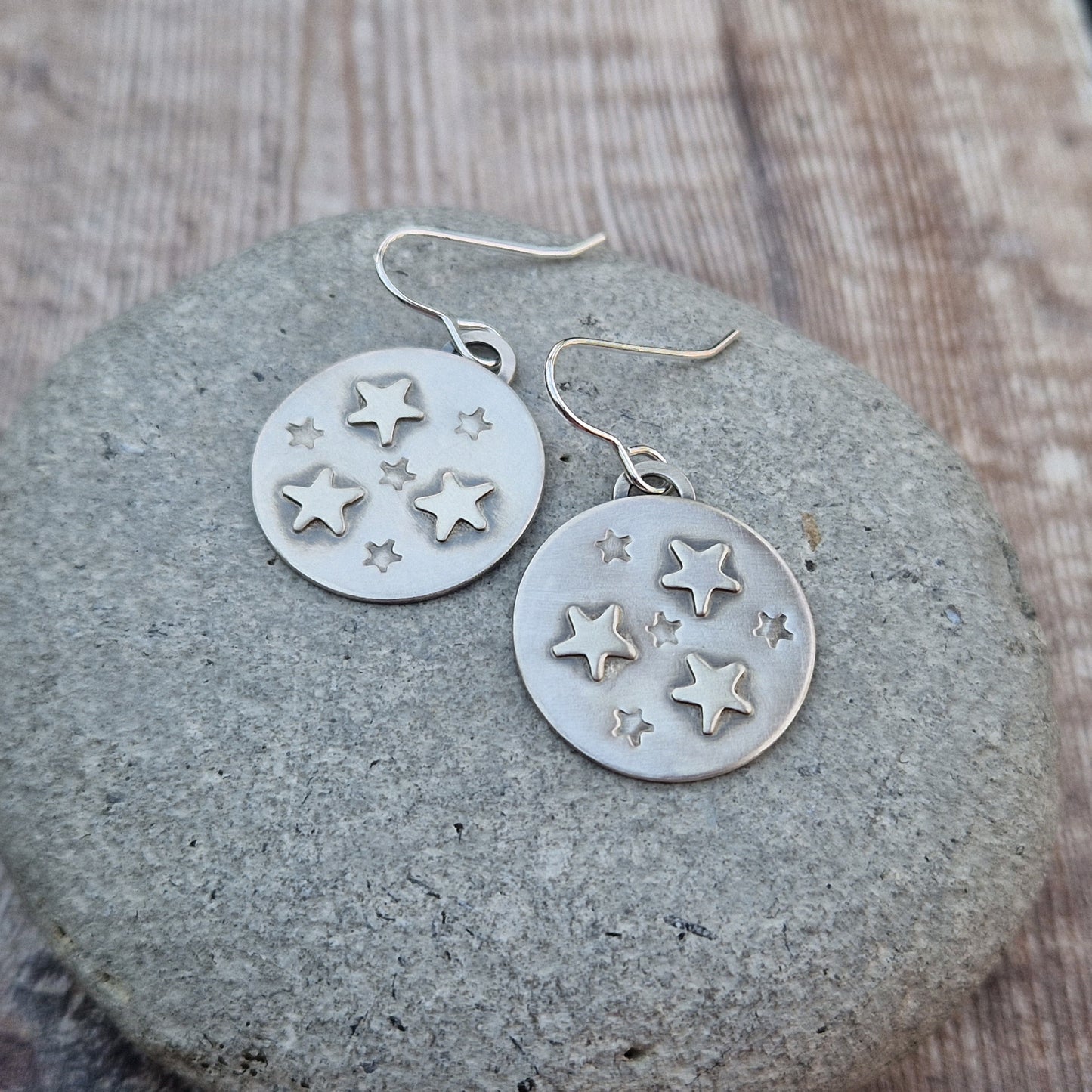 Each earring is an oxidised silver disc shape with three raised five-pointed stars and four indented smaller five-pointed stars in a random pattern on each disc. The oxidisation helps the star shapes stand out as if in shadow. Suspended from silver earring wire.