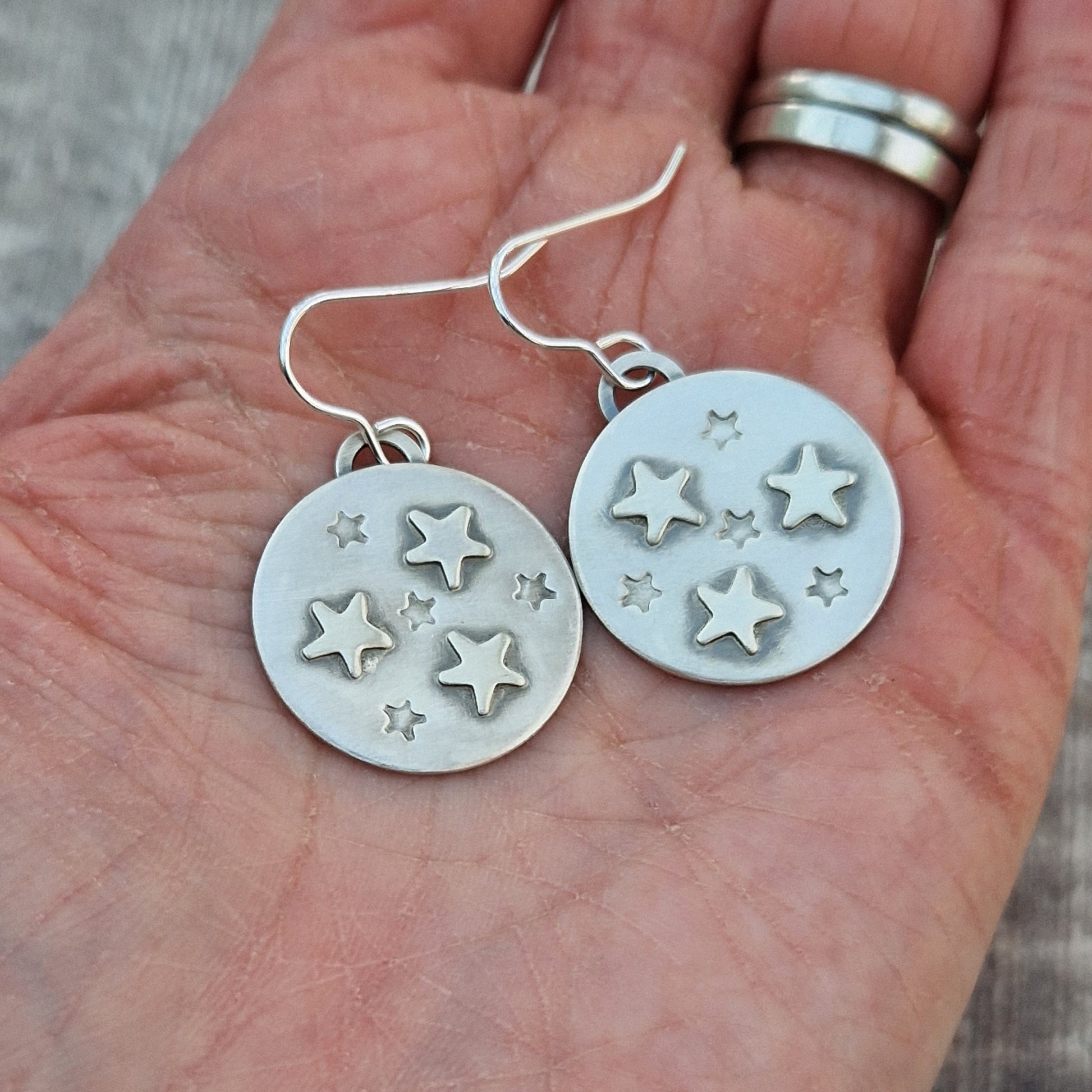 Each earring is an oxidised silver disc shape with three raised five-pointed stars and four indented smaller five-pointed stars in a random pattern on each disc. The oxidisation helps the star shapes stand out as if in shadow. Suspended from silver earring wire.